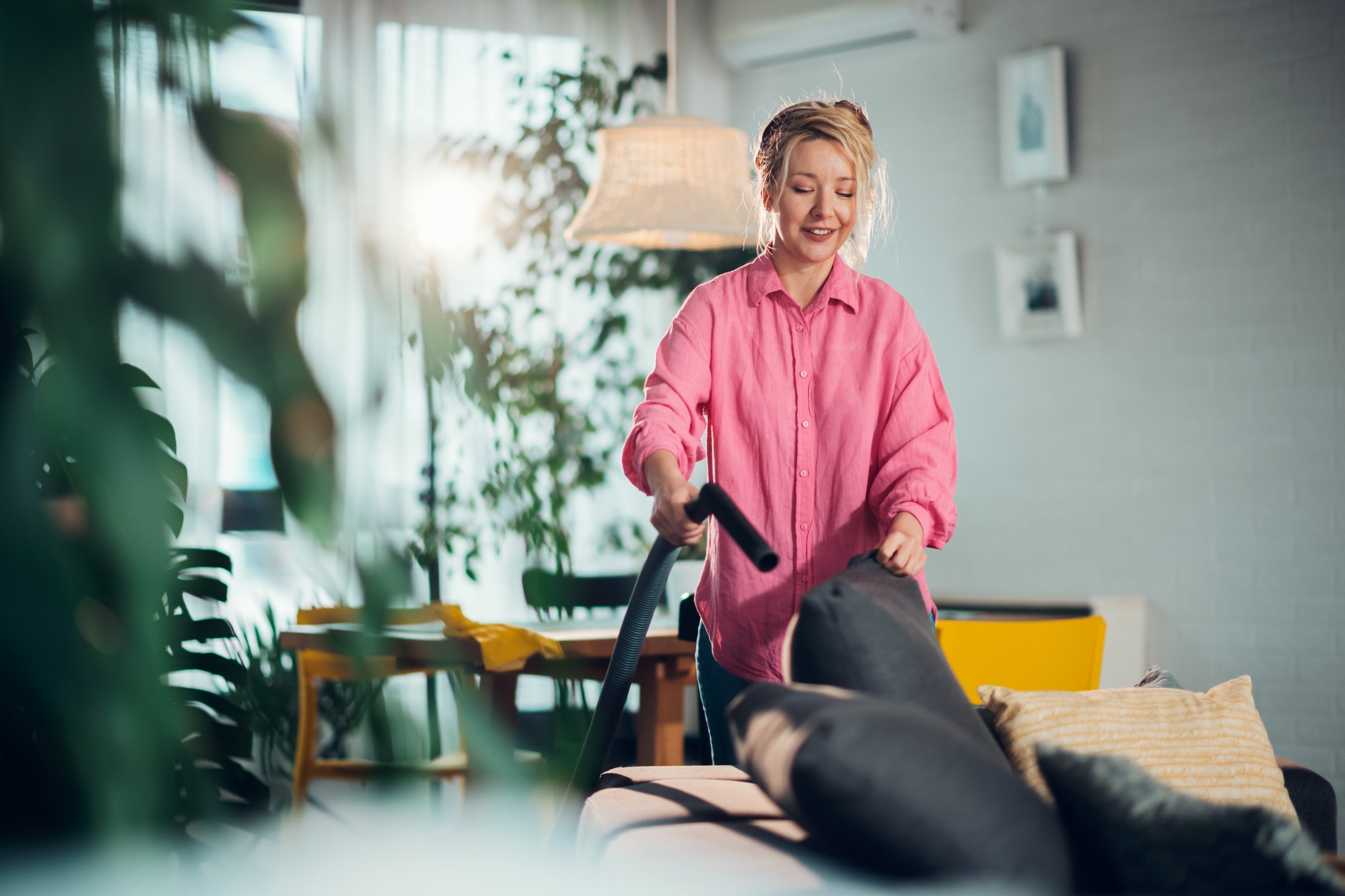 Smiling blonde woman vacuuming sofa pillows in bright living room with many houseplants, enjoying her daily chores and keeping her home clean and tidy