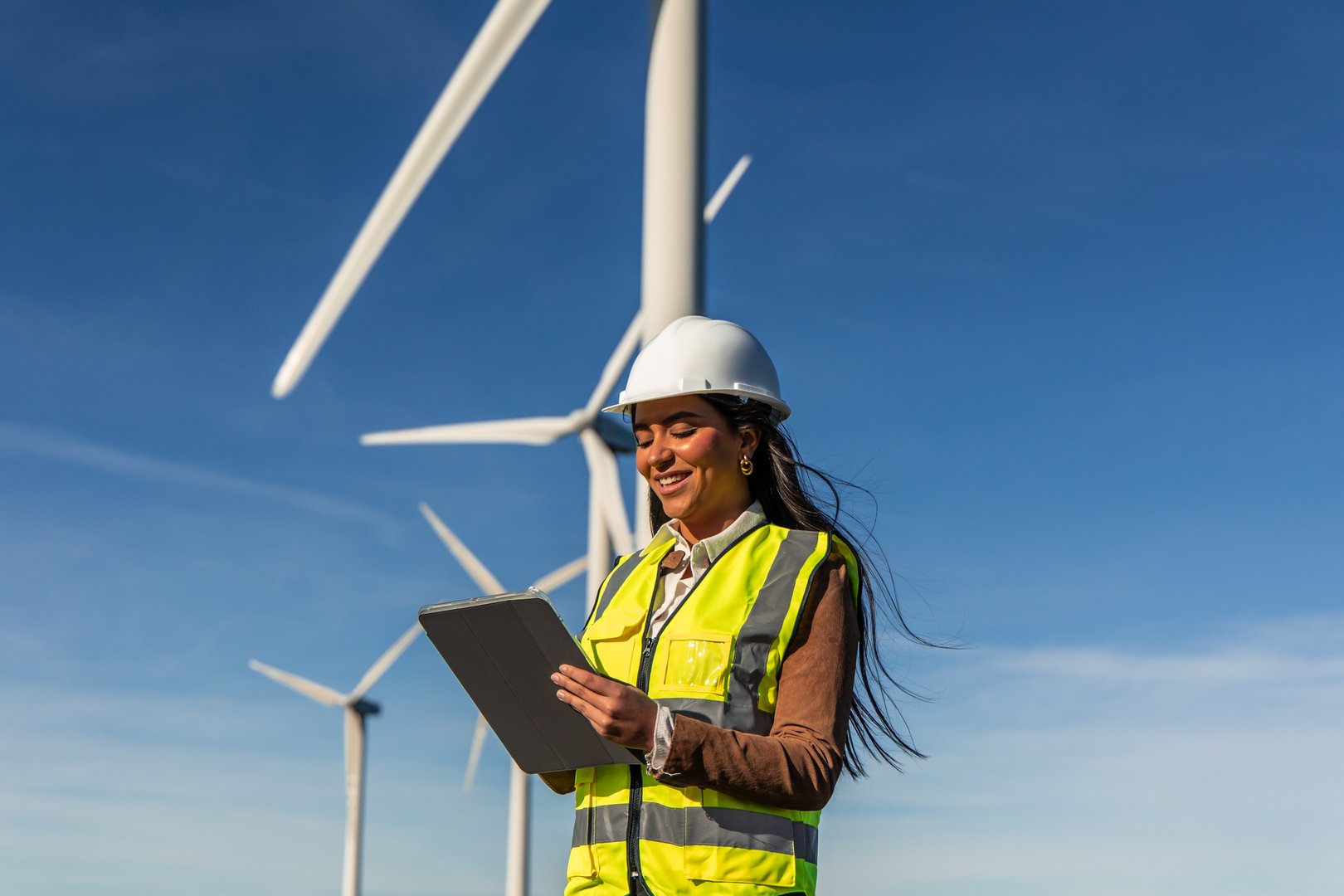 Smiling latin american female engineer wearing safety vest and helmet using digital tablet while inspecting wind turbines in a wind farm with blue sky in background