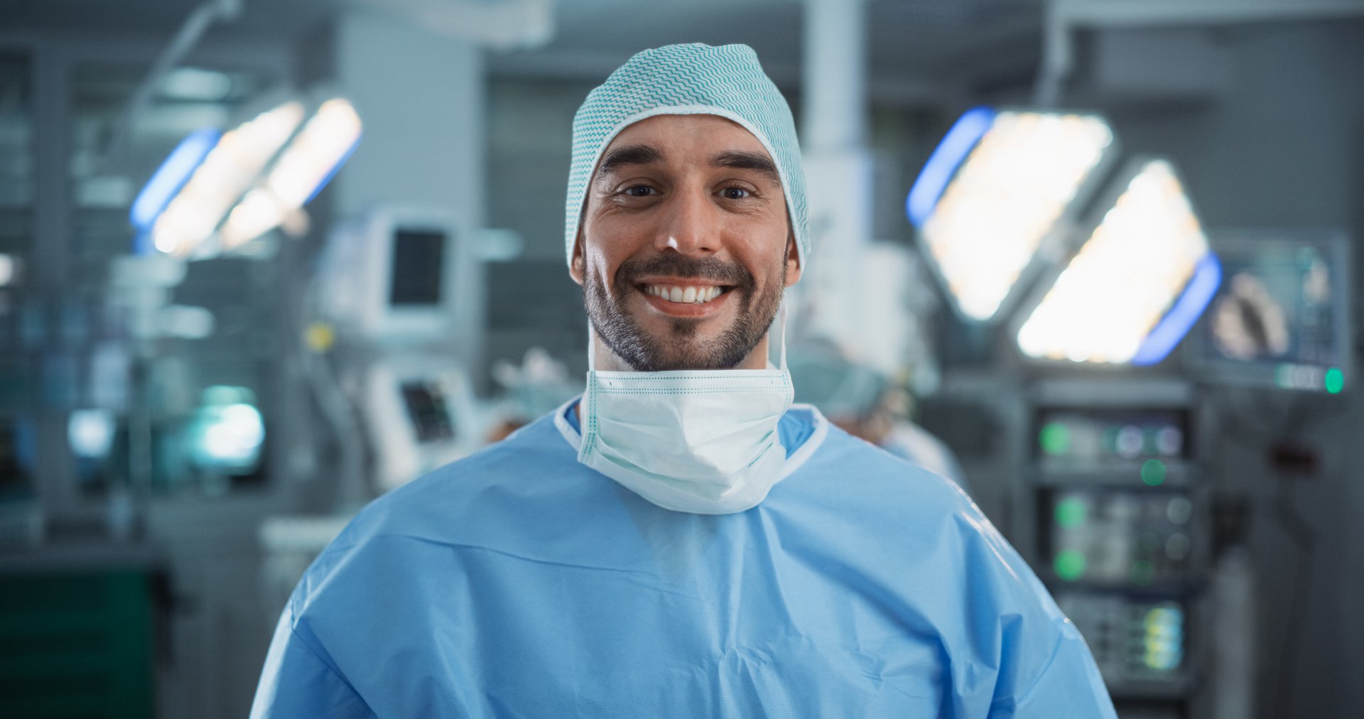 Portrait of a Caucasian Surgeon Taking off His Surgical Mask After a Successful Operation. Healthcare Professional Posing, Looking at Camera and Smiling. Photo in a Modern Hospital Operating Room