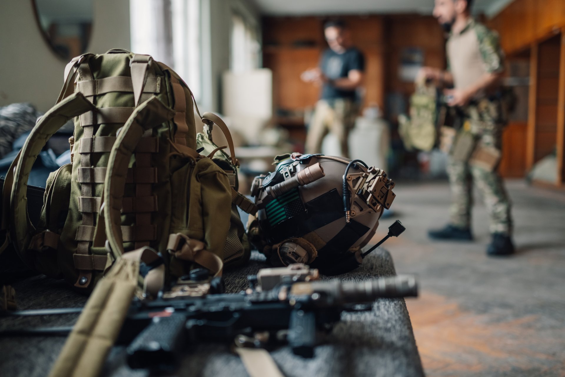 Close up of airsoft and tactical gear and equipment with players in camouflage uniforms in background. Selective focus on airsoft and tactical equipment with fighters getting ready for war simulation.