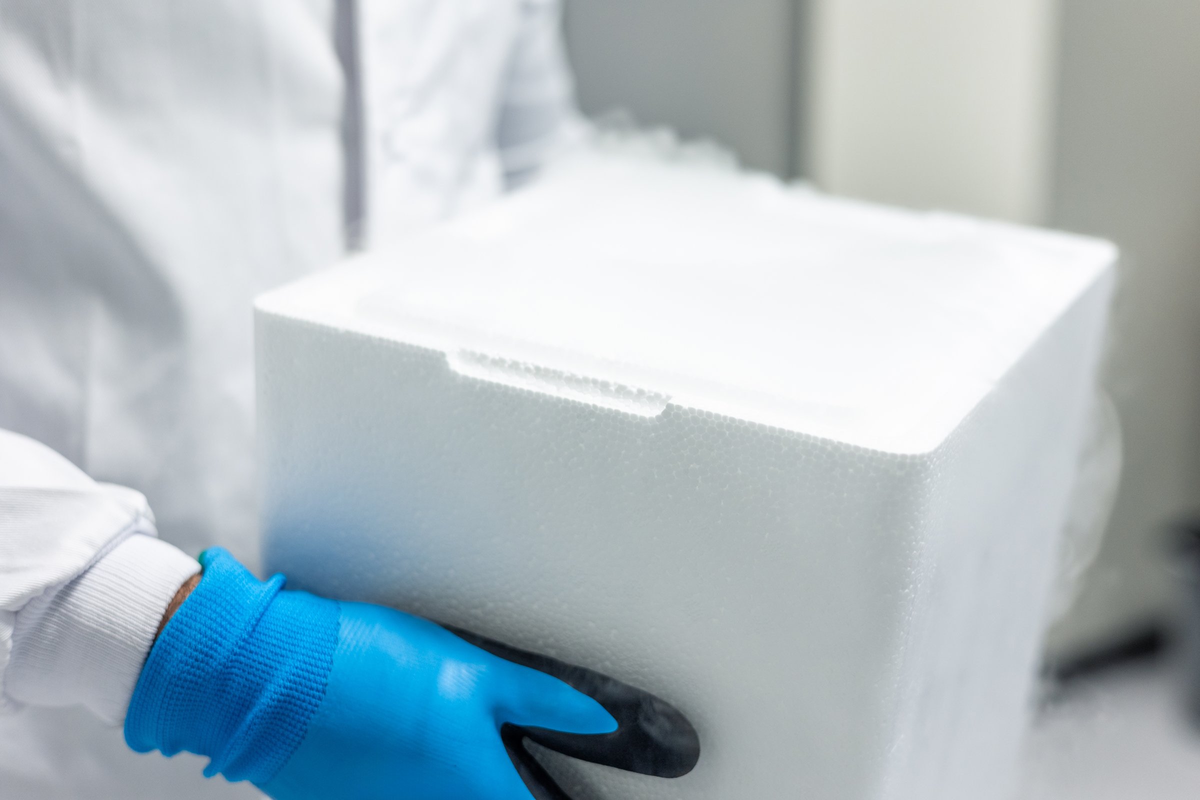 Scientist wearing a lab coat and gloves carefully carries a styrofoam box containing dry ice, emitting a white fog, in a scientific laboratory environment