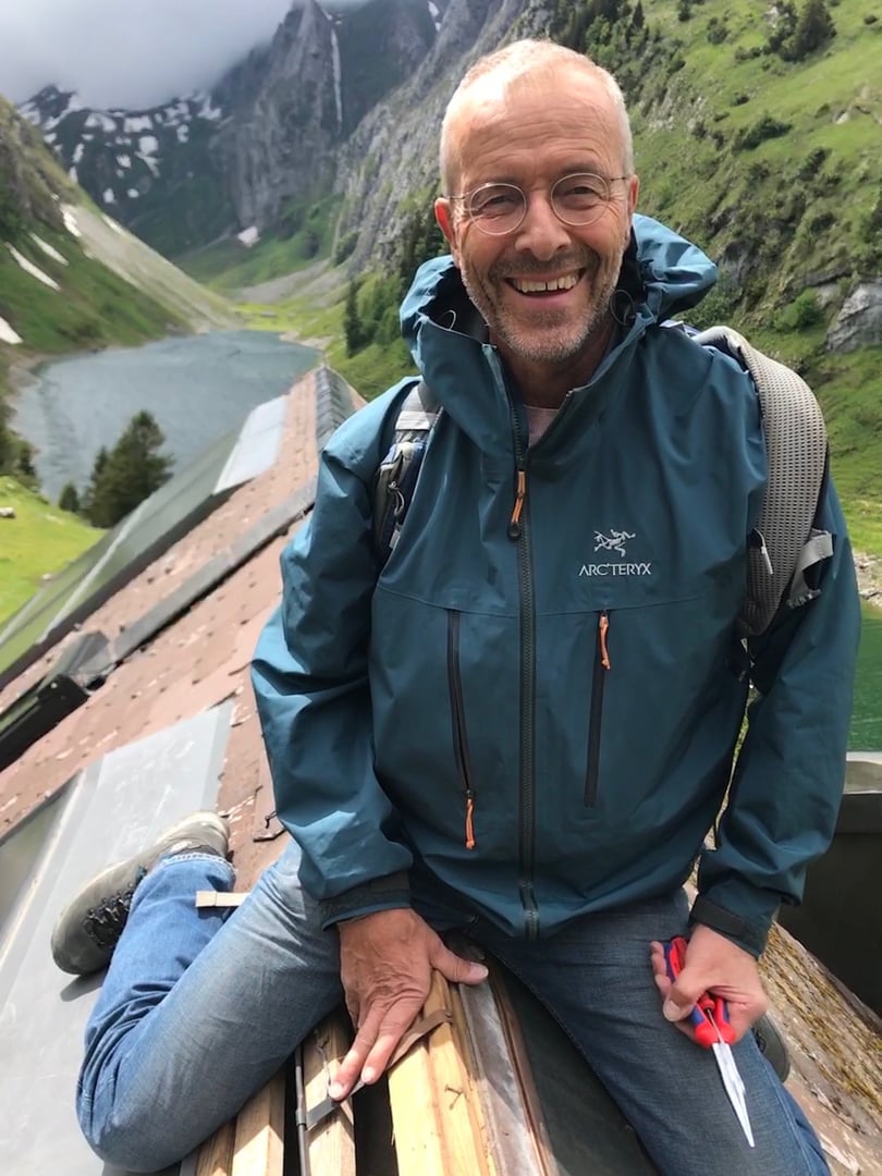 Smiling man in a blue jacket and jeans sitting on a wooden structure with a lake and mountains in the background.