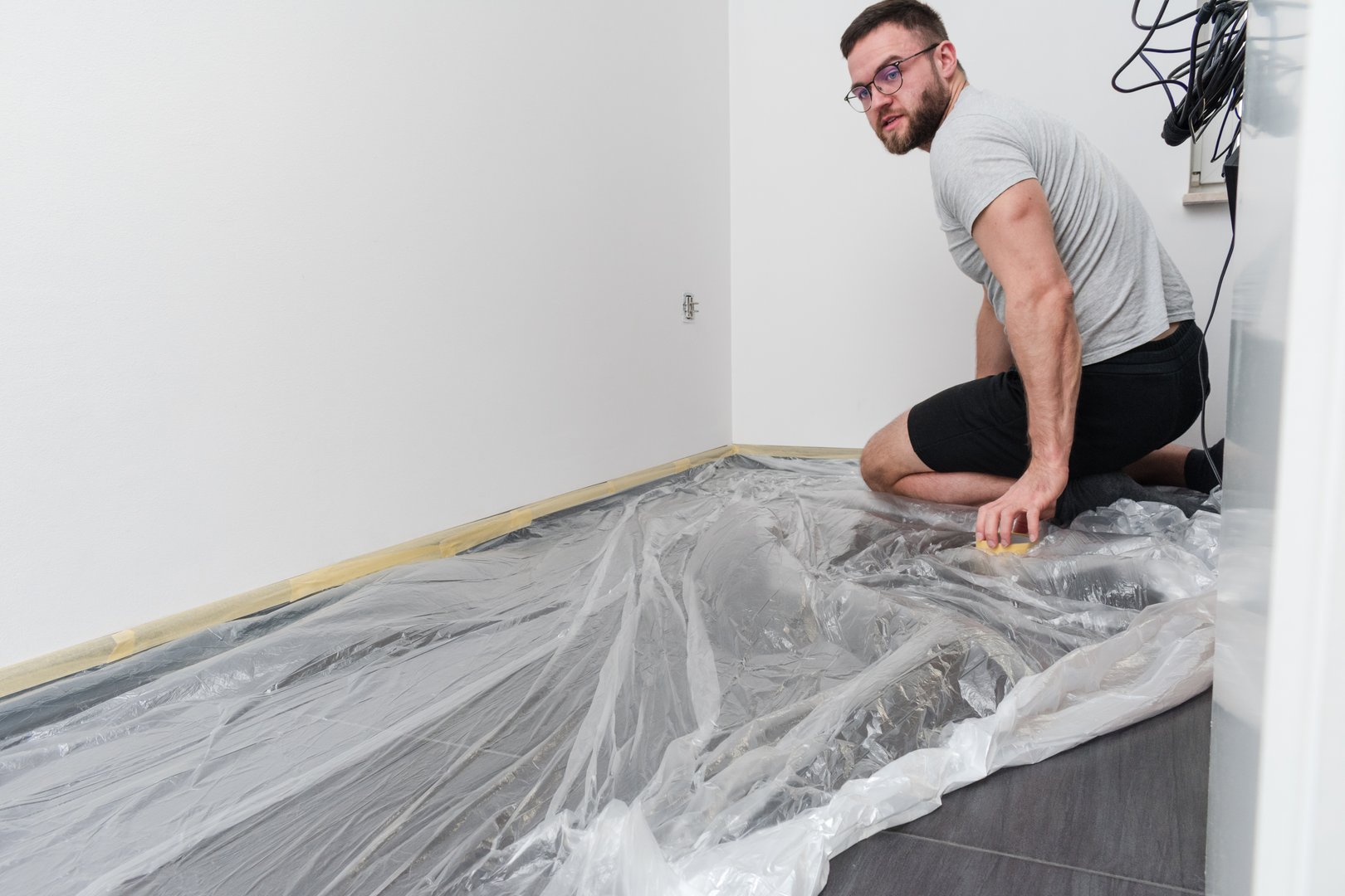 Man is kneeling on a floor with a plastic sheet on it. He is using a yellow tape to stick sheet to the floor