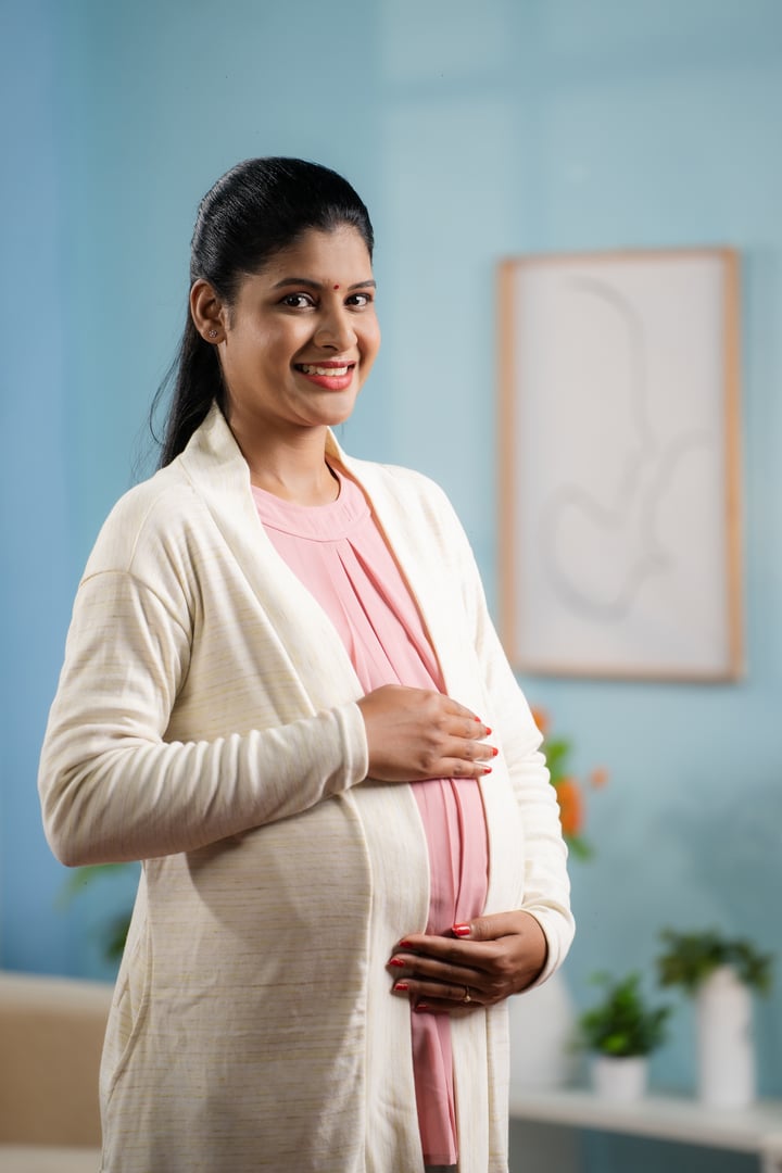 Verticak Portrait shot of happy smiling Indian Pregnant woman standing by holding tummy while looking camera - concept of motherhood, expecting baby and parenthoodVertical Portrait shot of happy smiling Indian Pregnant woman standing by holding tummy while looking camera - concept of motherhood, expecting baby and parenthood