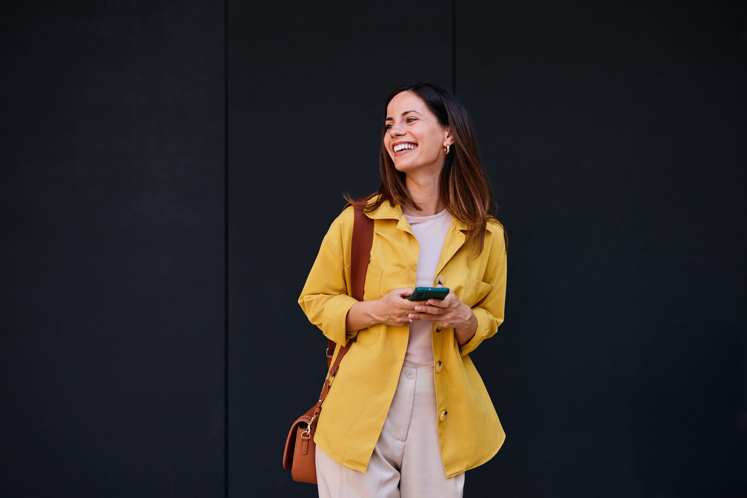 Cheerful woman holding a phone outdoors, dressed in a vibrant jacket, looking happy.