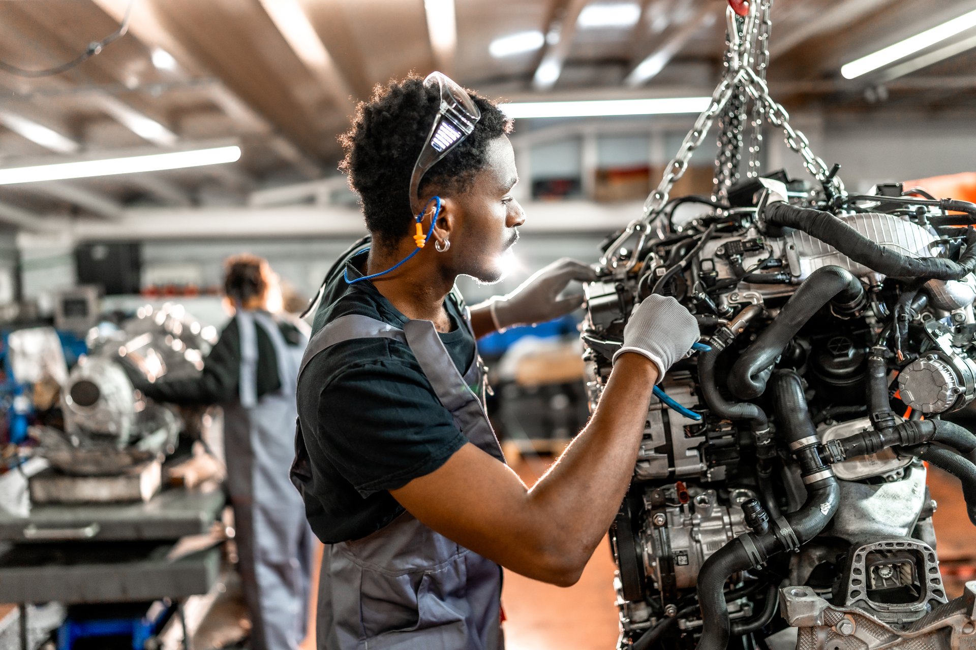 Young mechanic repairing a car engine hanging from a chain hoist, wearing safety goggles and earplugs, in a professional workshop