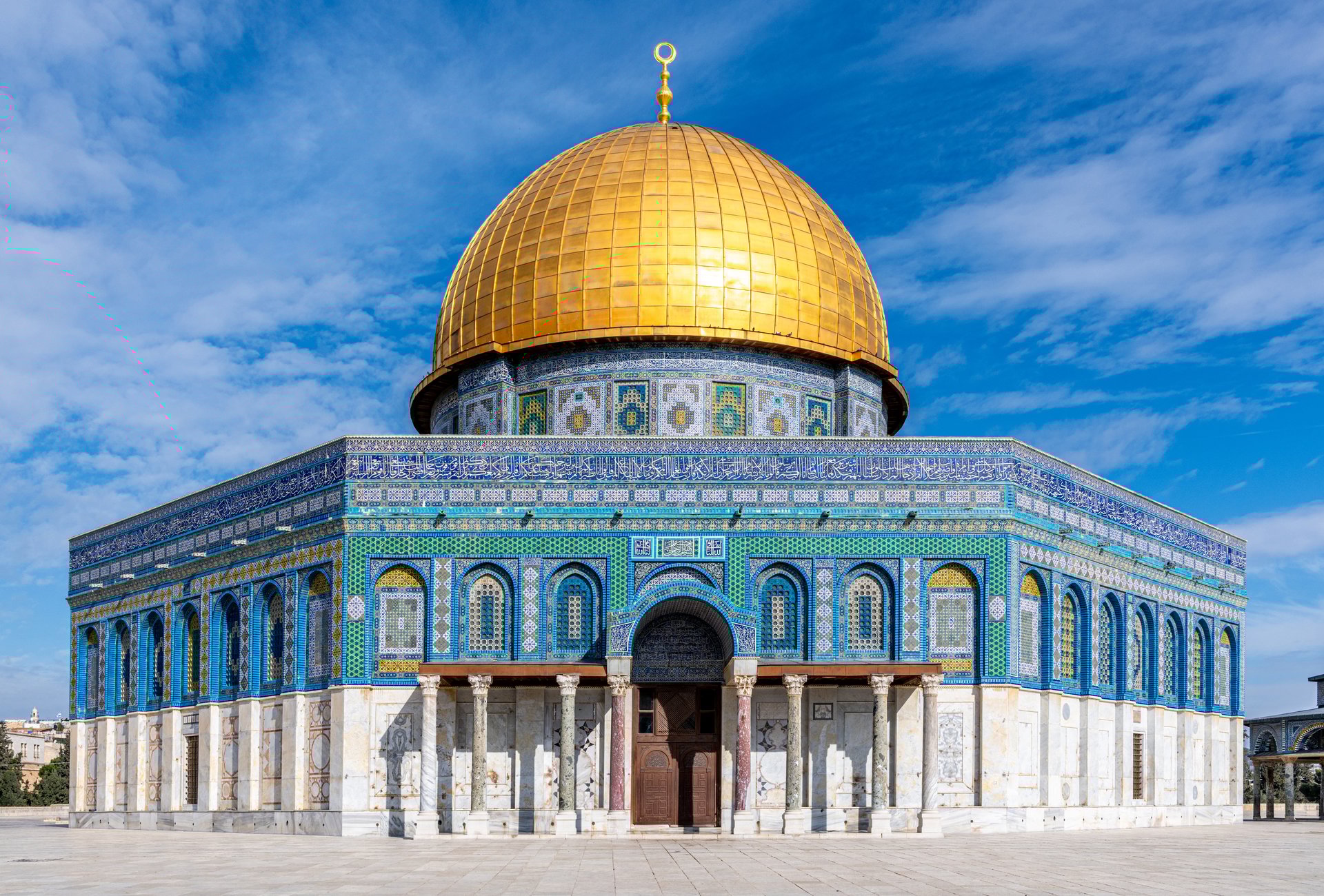 The Dome of the Rock in Jerusalem's Old City features a large, gleaming golden dome atop intricate blue, green, and white geometric tile patterns. Arabic calligraphy adorns the facade, adding to its intricate design. The building's architecture reflects Islamic artistry, with arches and detailed ornamentation. The dome and vibrant tiles create a striking contrast against the clear blue sky. This site holds significant historical and religious importance.
