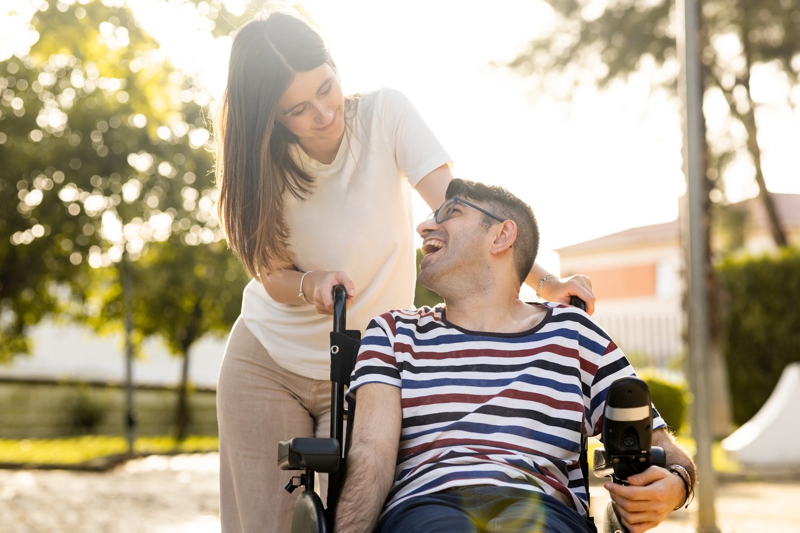 An adult man with a disability sits in an automatic wheelchair with a young woman in a park at sunset.The woman and the man look at each other happily. Concept of support for disabled people.