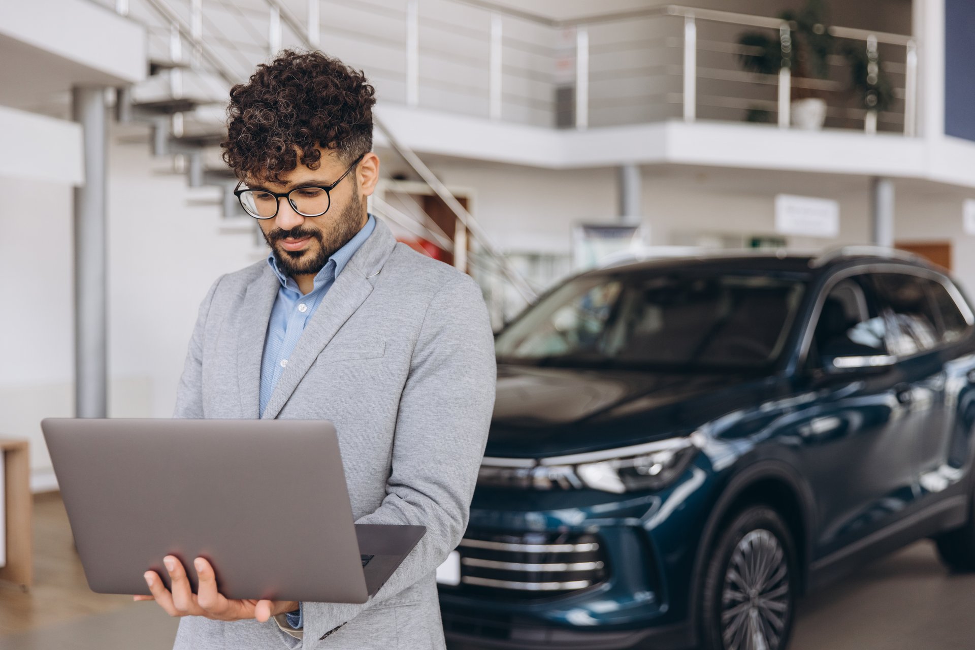 Salesman using laptop in car dealership showroom, consulting data for selling new car to customer