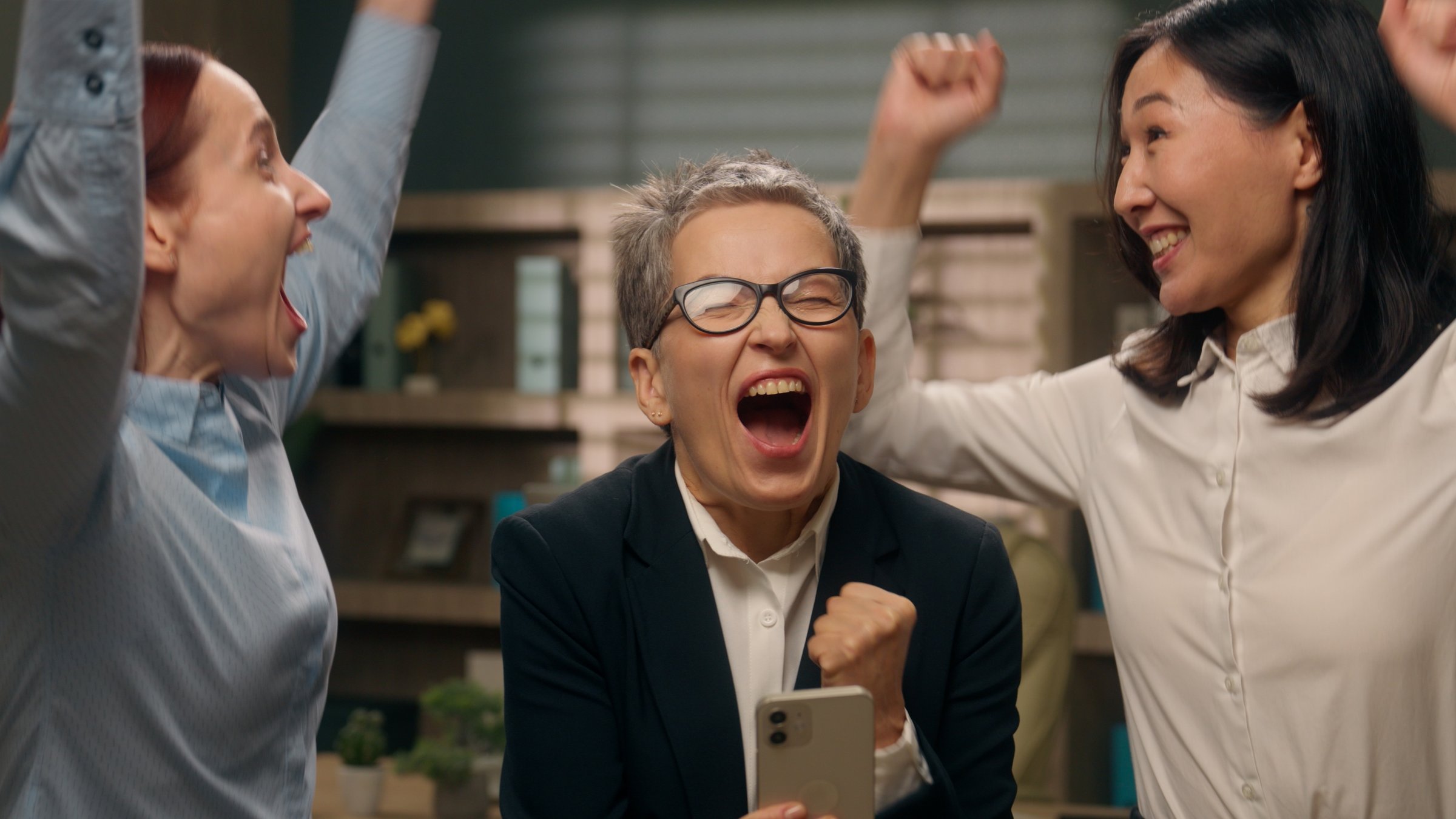 Happy excited women colleagues team group holding mobile phone multinational read good news businesswomen winning online finance celebrating office success offer ladies bet multiracial business people. High quality 4k footage