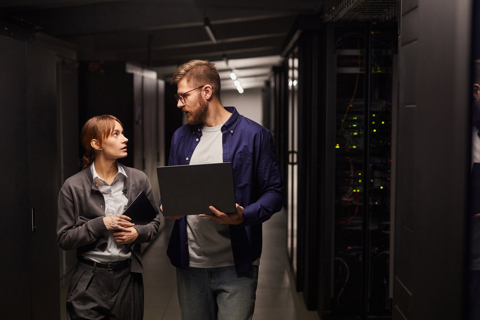 Two IT professionals discussing tasks while holding equipment in data center server room, surrounded by servers and technology