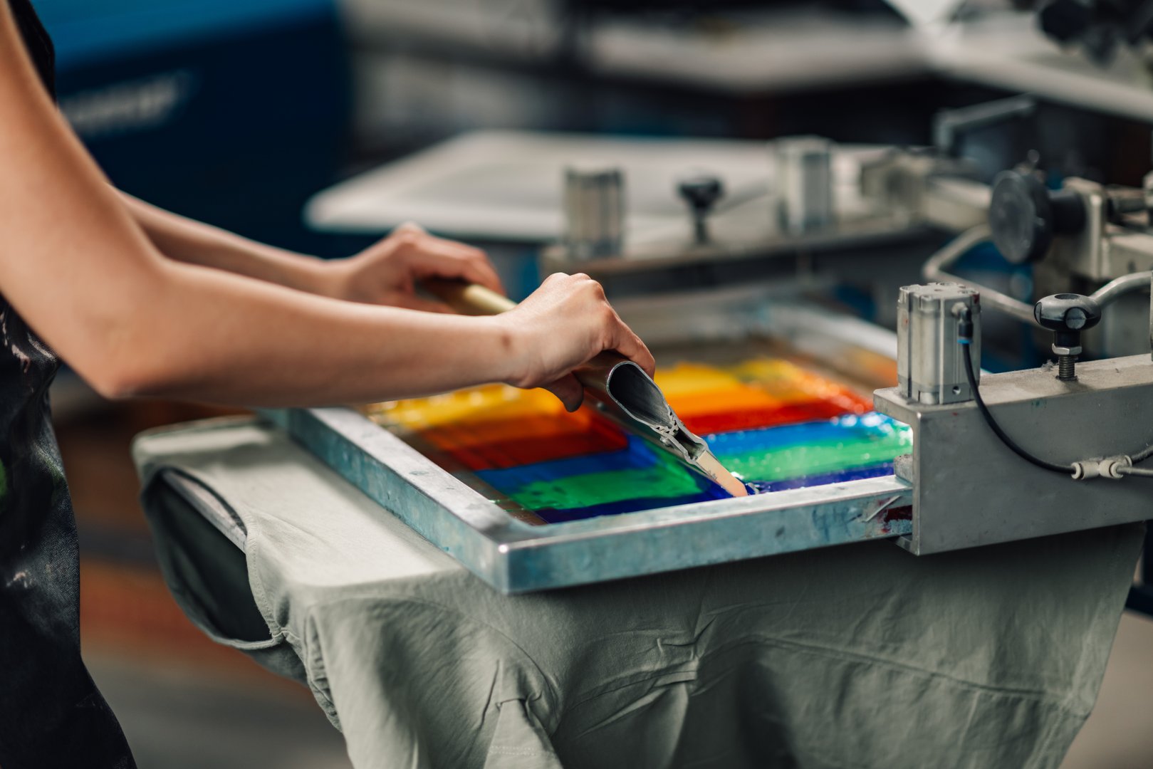 Close up of graphic technician's hands silkscreen printing on textile products and t-shirts with squeegee at printing workshop.