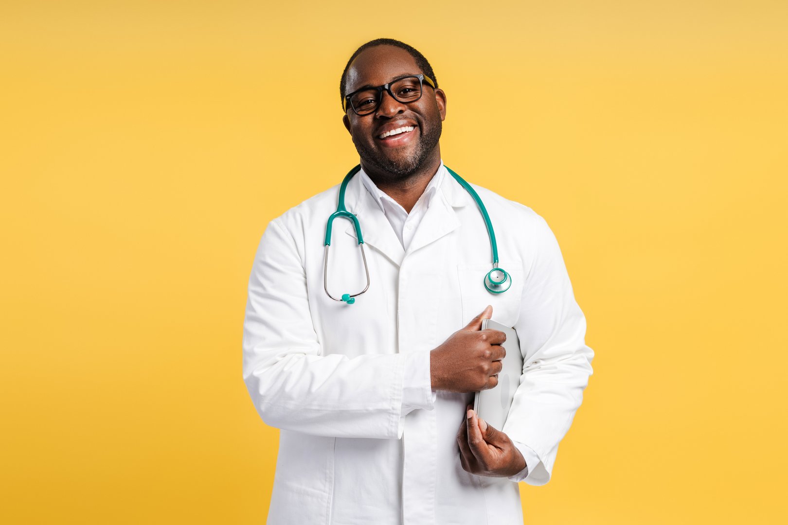 Portrait of happy African American doctor wearing white coat with stethoscope posing with laptop, isolated in yellow background