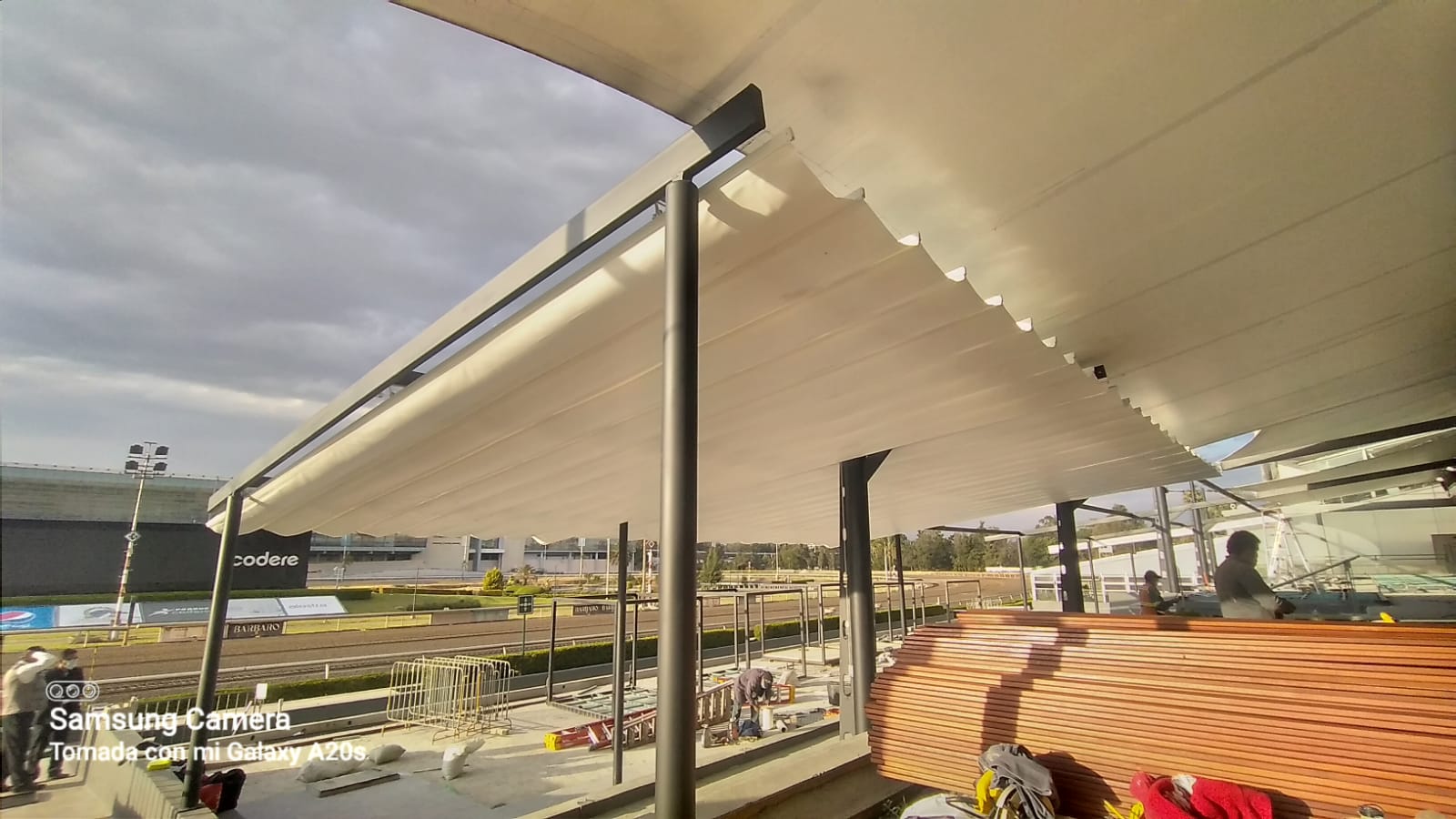 A large outdoor canopy structure with a wavy design, supported by metal beams. Construction materials are visible below.