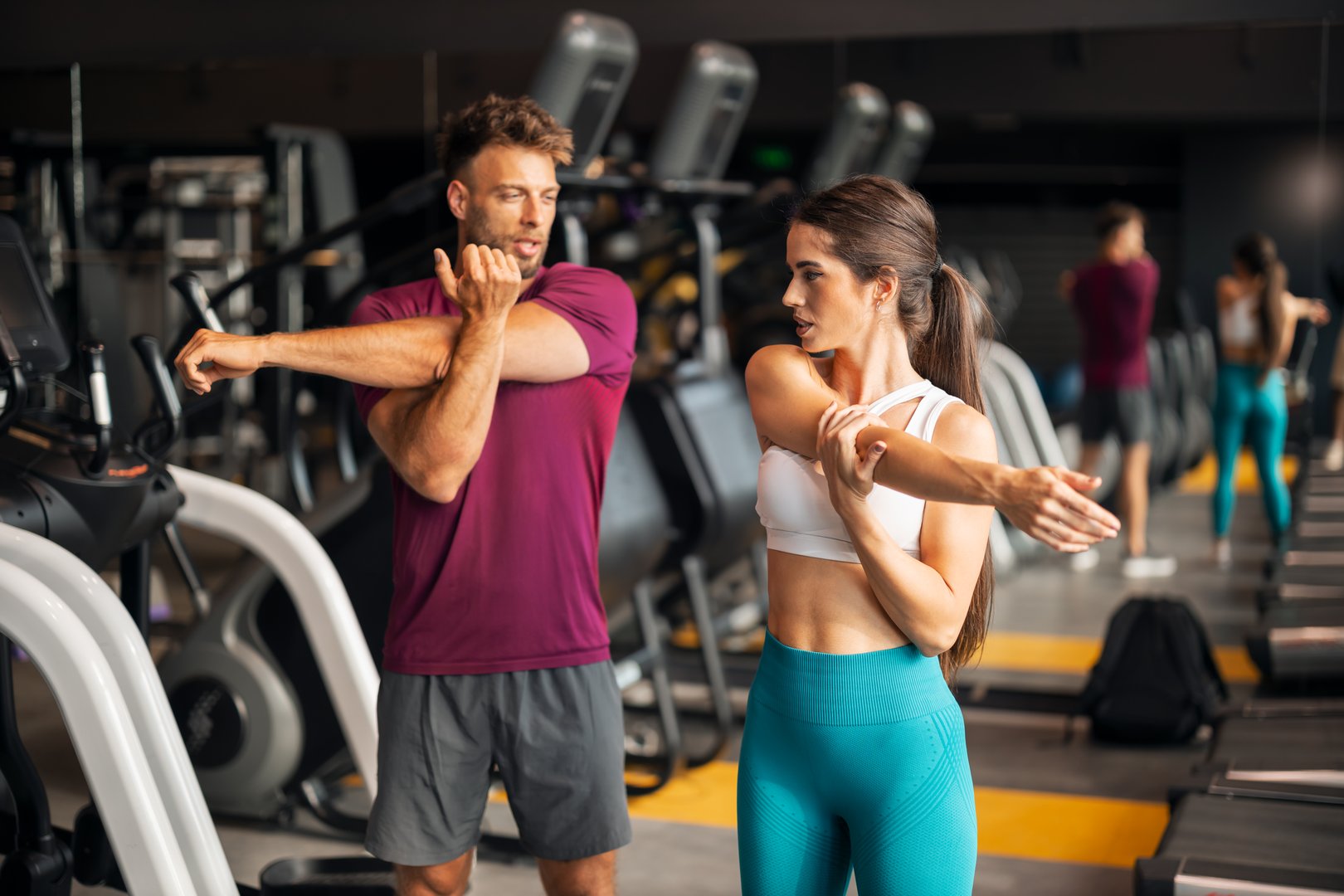 A fit young adult man and woman warming up for their workout and stretching their arms at the gym while talking.