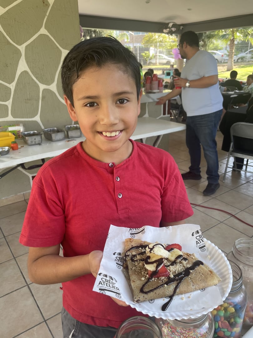 Boy in red shirt holding a dessert crepe with chocolate sauce and strawberries, smiling at an outdoor event.