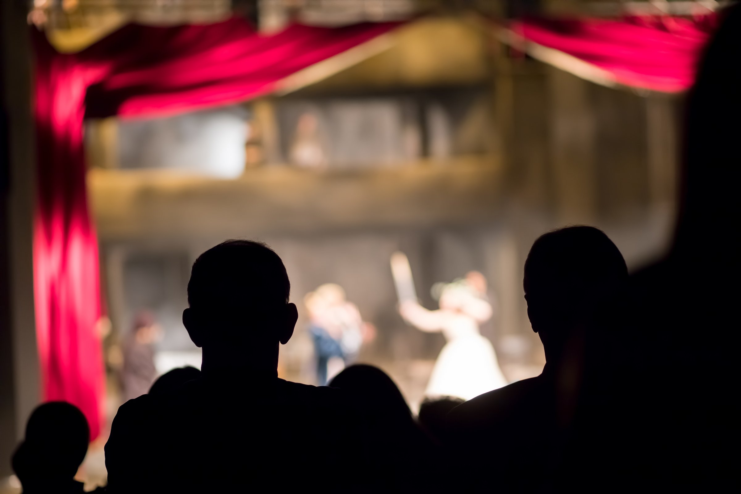 Silhouettes of audience members watching a ballet performance on stage with red curtains and dim lighting.