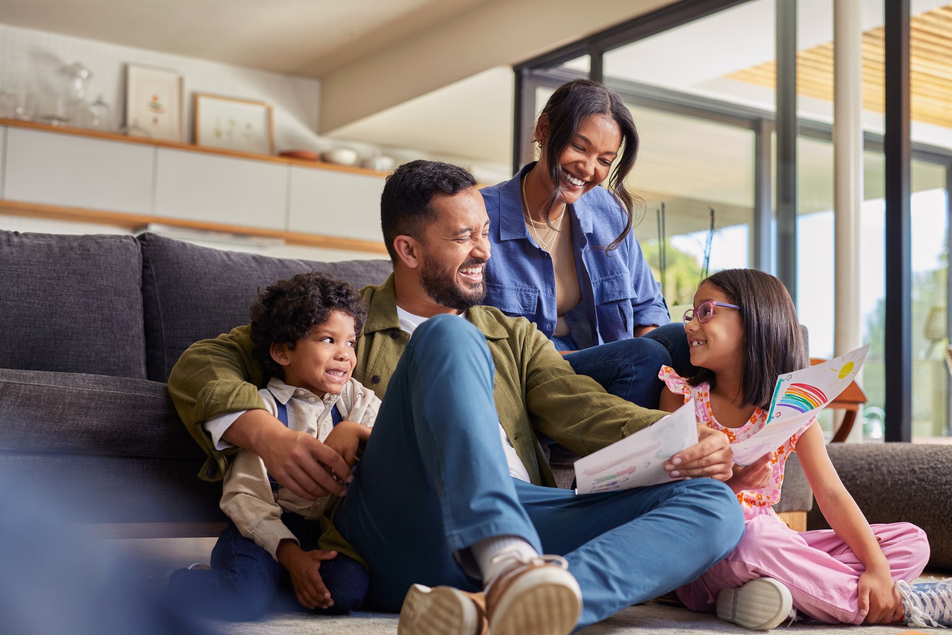 Creative happy smiling daughter showing artwork to family in living room. Proud little artist sharing her drawings with family at home. Little cute girl presents a hand drawn papers to her parents as they share a tender moment on a couch.
