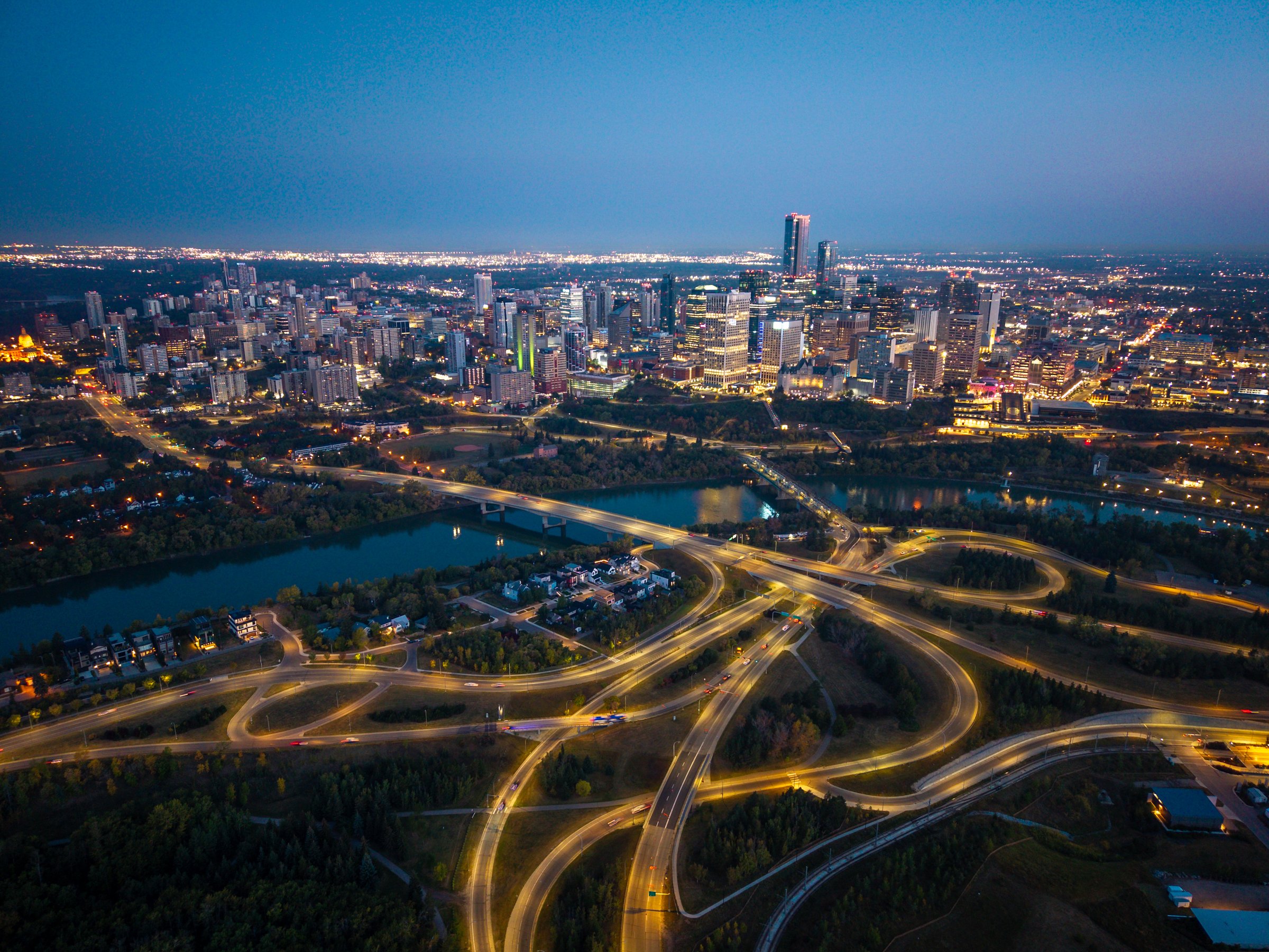 Aerial photograph of downtown Edmonton, Alberta at night, showcasing the illuminated city skyline, highways, and bridges over the North Saskatchewan River. The glowing road network and traffic patterns highlight the modern infrastructure and urban energy of Alberta's capital city. This image captures themes of transportation, development, technology, and Canadian city life.