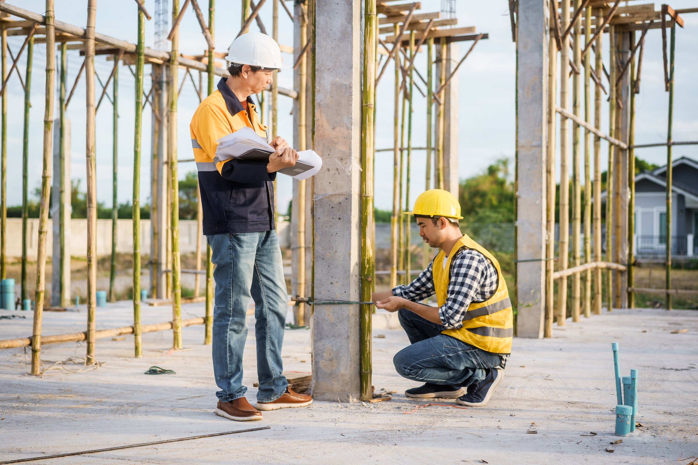 An elderly man and a middle-aged Asian man, both wearing hard hats and reflective vests, closely inspect construction blueprints on site amid an active construction site with beams and columns