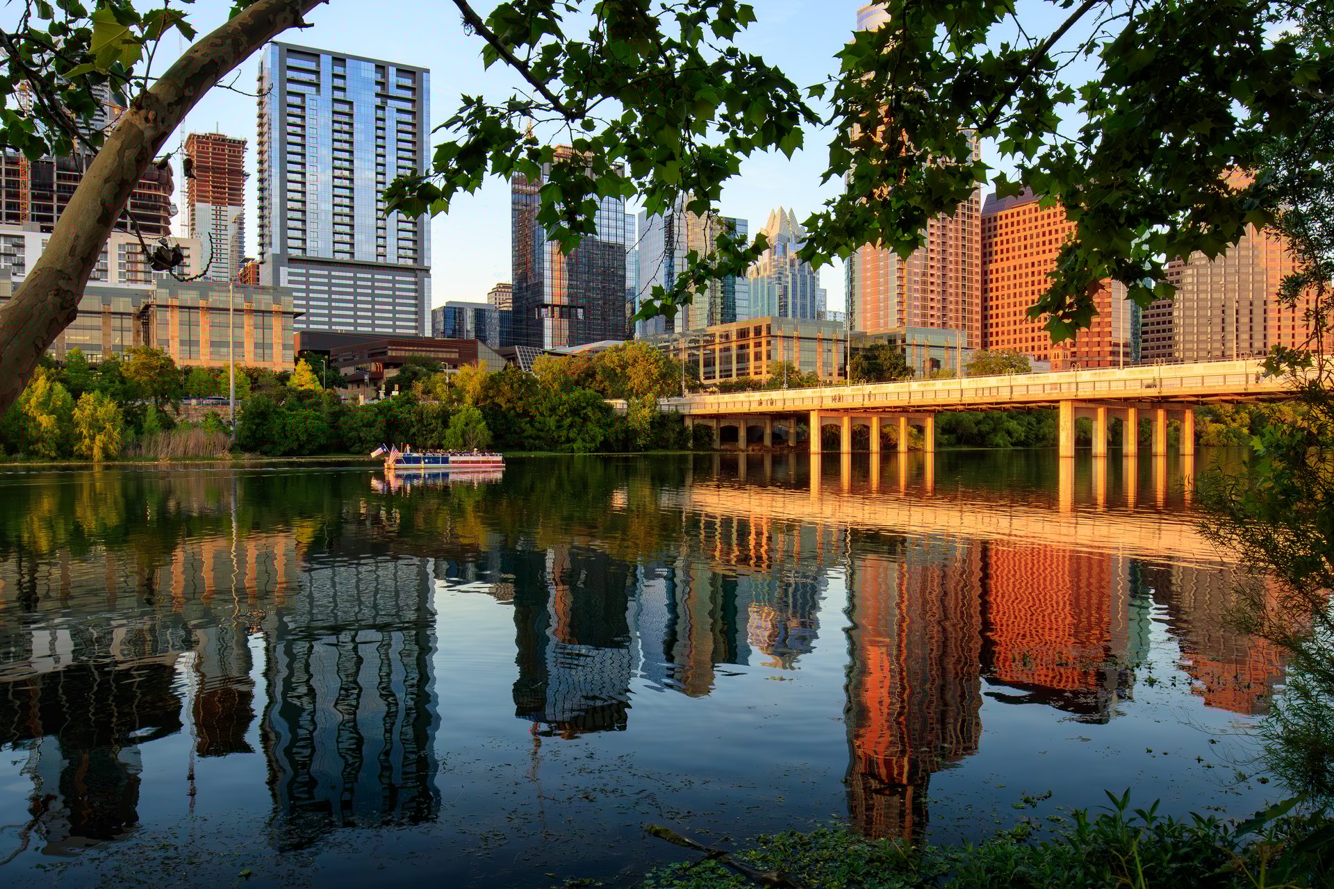 A serene body of water, Lady Bird Lake, reflects the skyline of the modern city of Austin Texas with a clear sky above. In the foreground, lush greenery frames the tranquil scene where a small boat with people can be seen on the water. Located at Auditorium Shores, Town Lake, Lady Bird Lake Metropolitan Park.