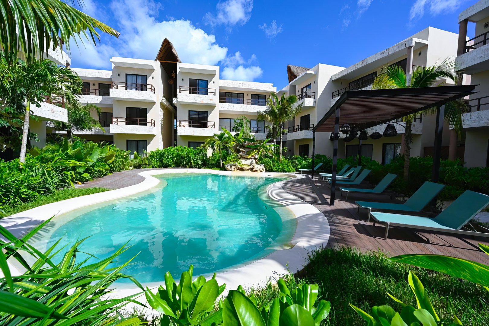 Tropical resort pool surrounded by lush greenery, sun loungers, and modern apartment buildings under a blue sky.