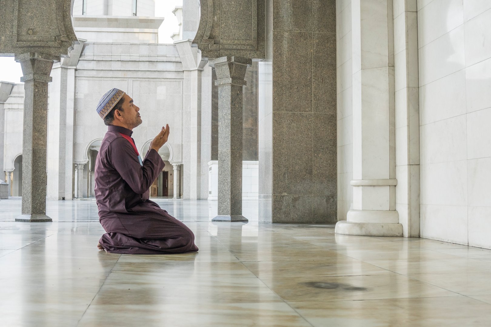 Man in traditional attire kneeling and praying inside a mosque with marble floors and tall stone columns.
