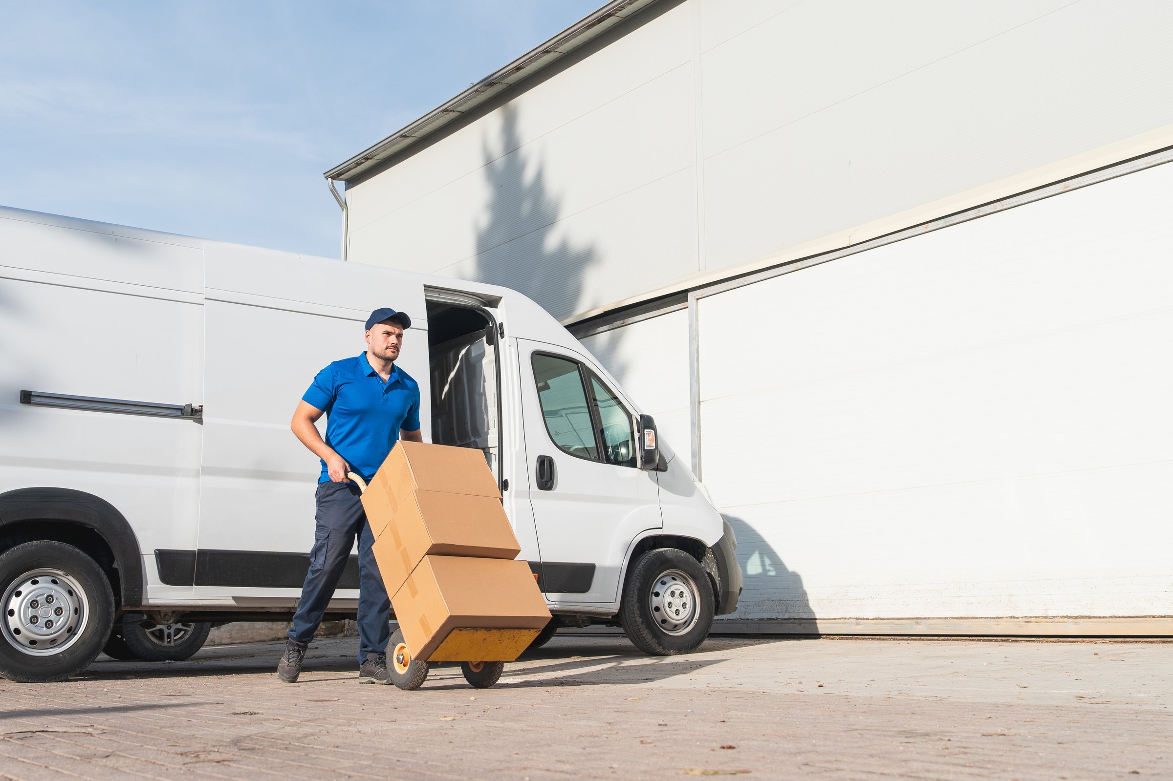 Delivery man in blue uniform transporting cardboard boxes on a hand truck outside a warehouse with a white van.