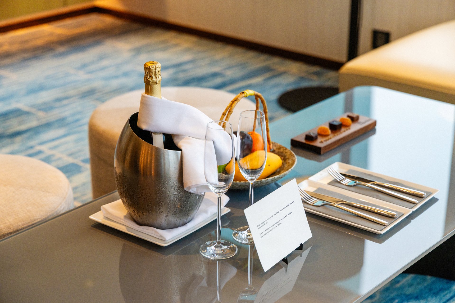 Luxury hotel room with champagne, fruit basket, and chocolate on glass table. Elegant dining setting with wine glasses, cutlery, relaxing atmosphere, upscale hospitality.