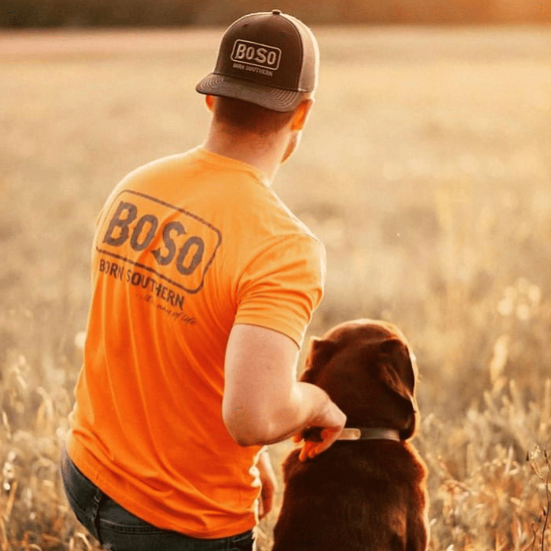 Person wearing an orange shirt and cap with BOSO logo, sitting with a brown dog in a field at sunset.