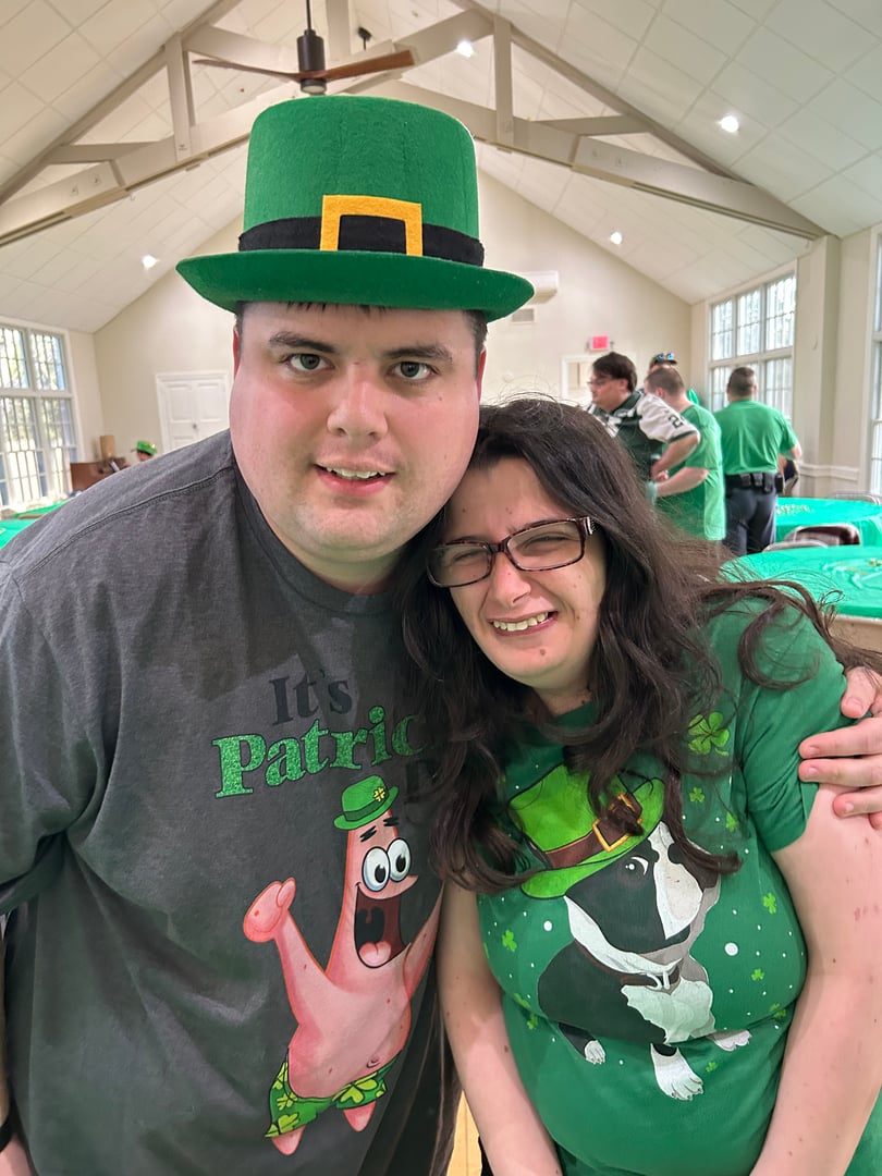 Two people wearing St. Patrick's Day-themed outfits stand indoors, smiling. One wears a green hat, the other a green T-shirt.