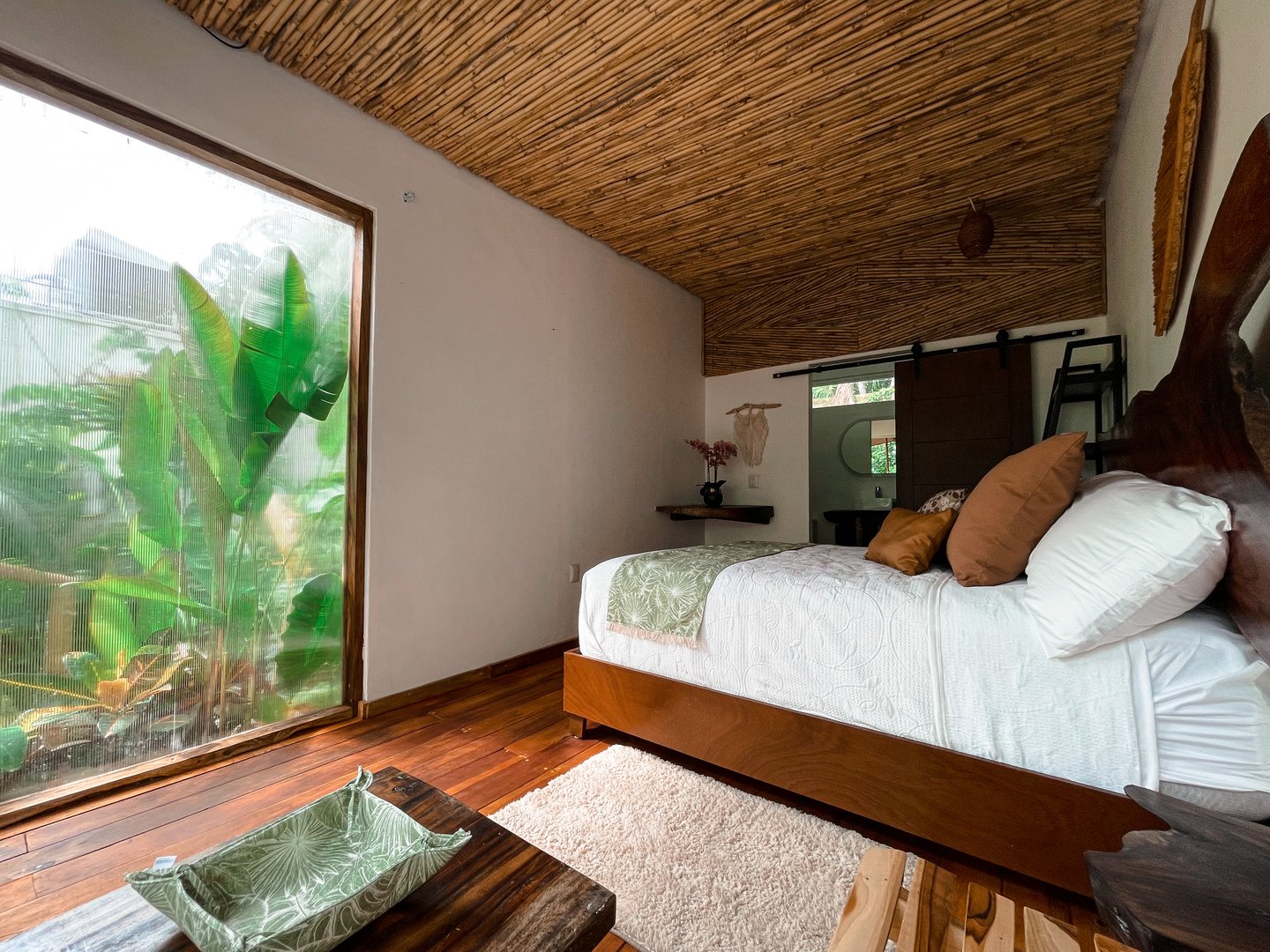 Rustic bedroom with wooden accents, large window with greenery view, and cozy bed with green and brown decor.