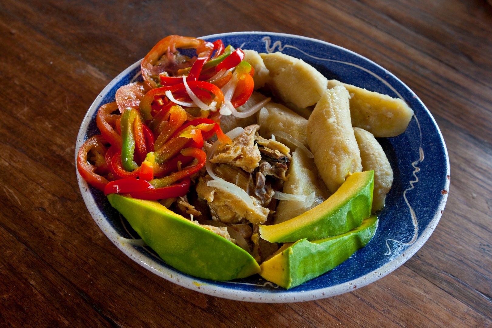A plate with cooked plantains, slices of avocado, and sautéed vegetables, including red and green peppers, on a wooden table.