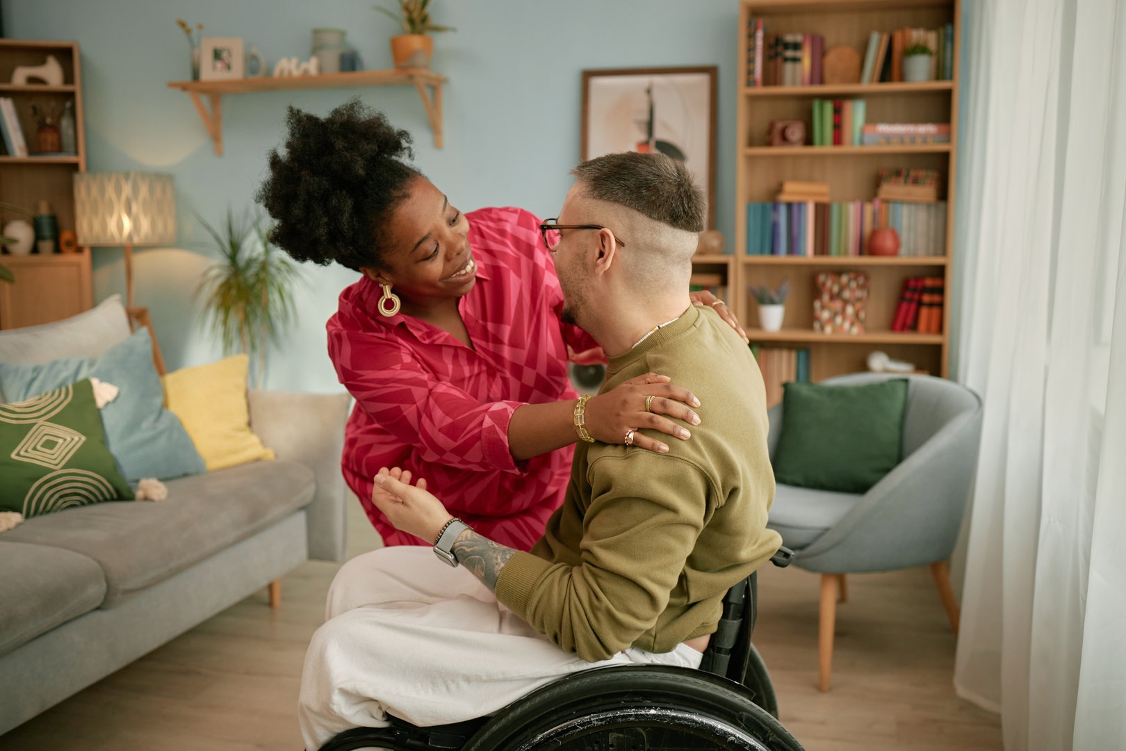 Black woman smiling and embracing Caucasian man with disability in wheelchair during birthday celebration at home, both appearing as young adults, sharing joyful moment together