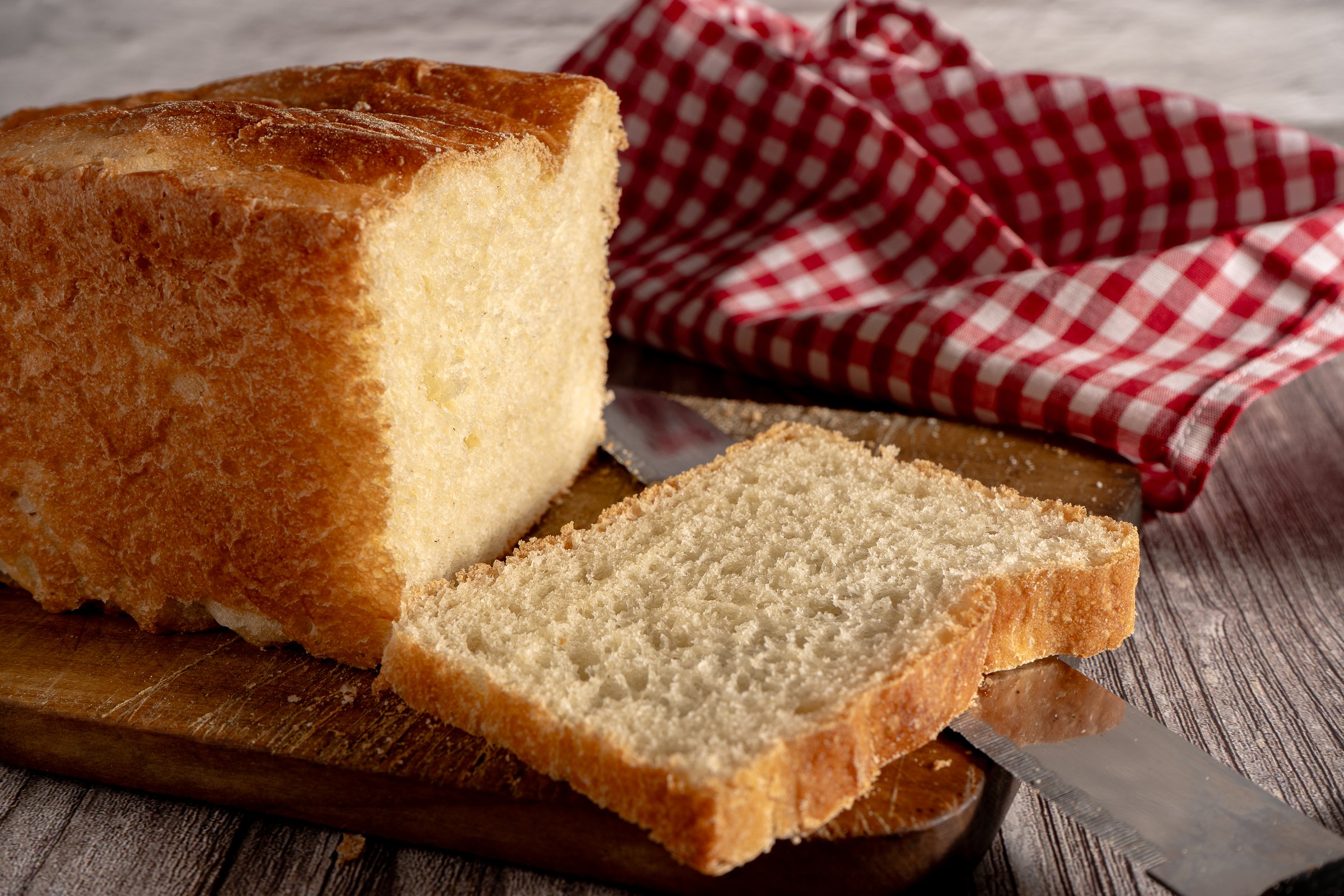 Freshly baked white bread loaf with a slice cut off, resting on a wooden cutting board with a bread knife and a red checkered napkin
