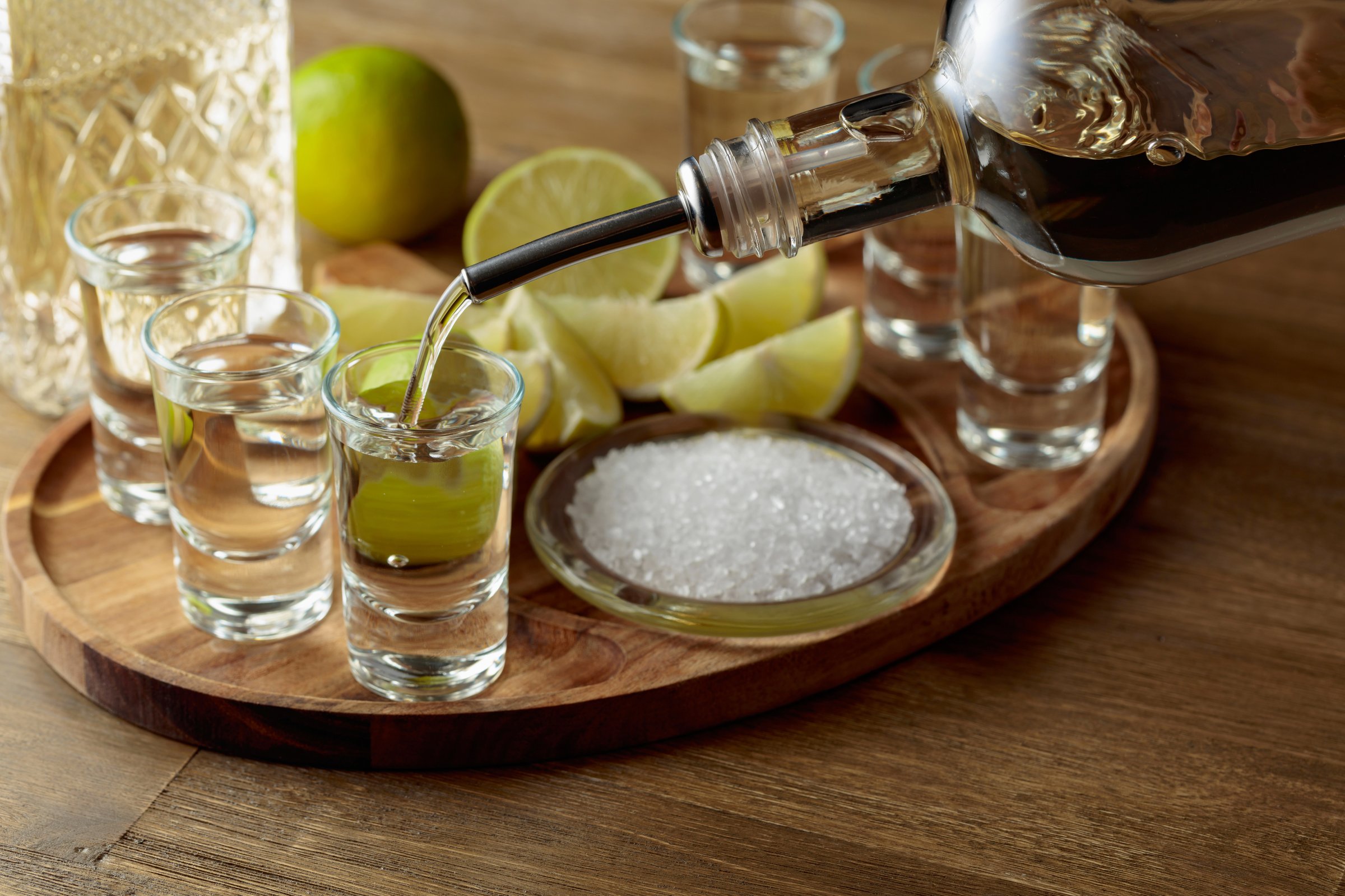 Gold tequila with salt and lime slices on a wooden tray. Tequila is poured into a shot.