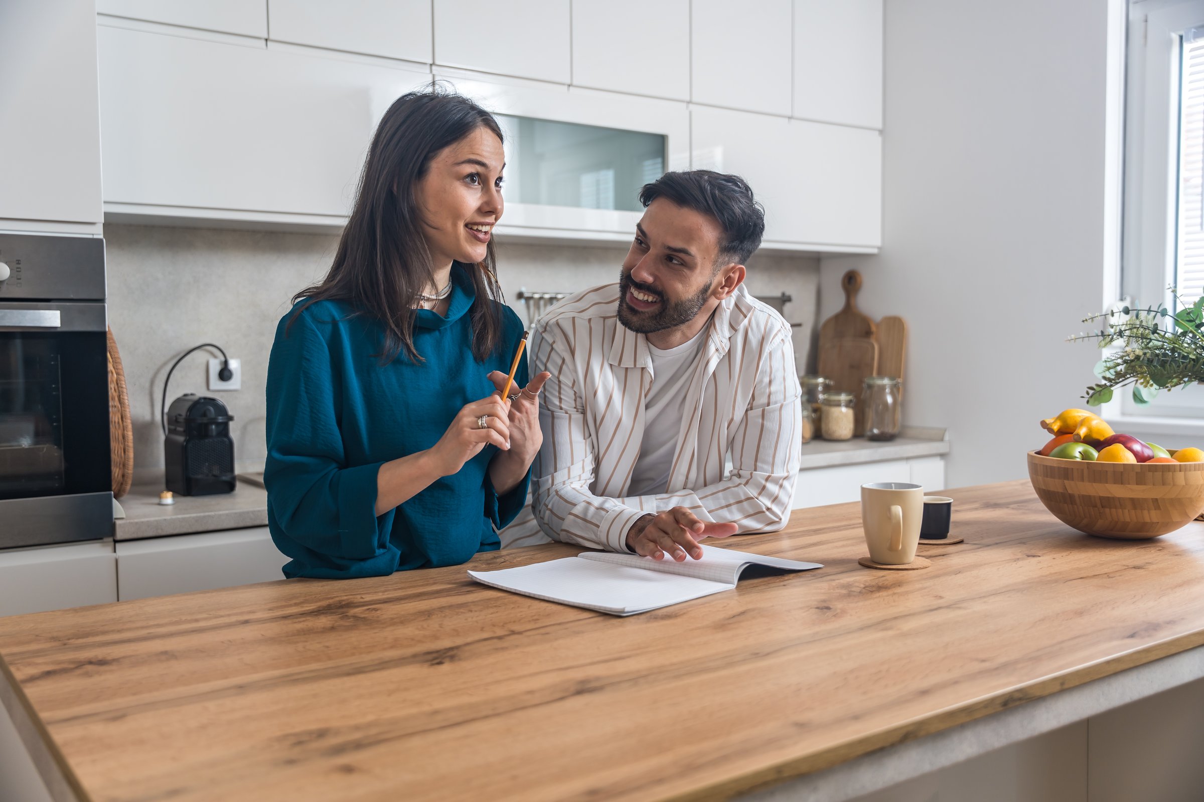 Young engaged couple standing together in their modern kitchen, writing wedding guest list and organizing catering menu, focused and smiling while preparing for their upcoming celebration at home