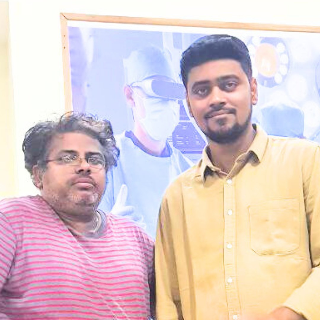 Two men posing indoors against a medical-themed background, one wearing a striped shirt, the other in a yellow shirt.