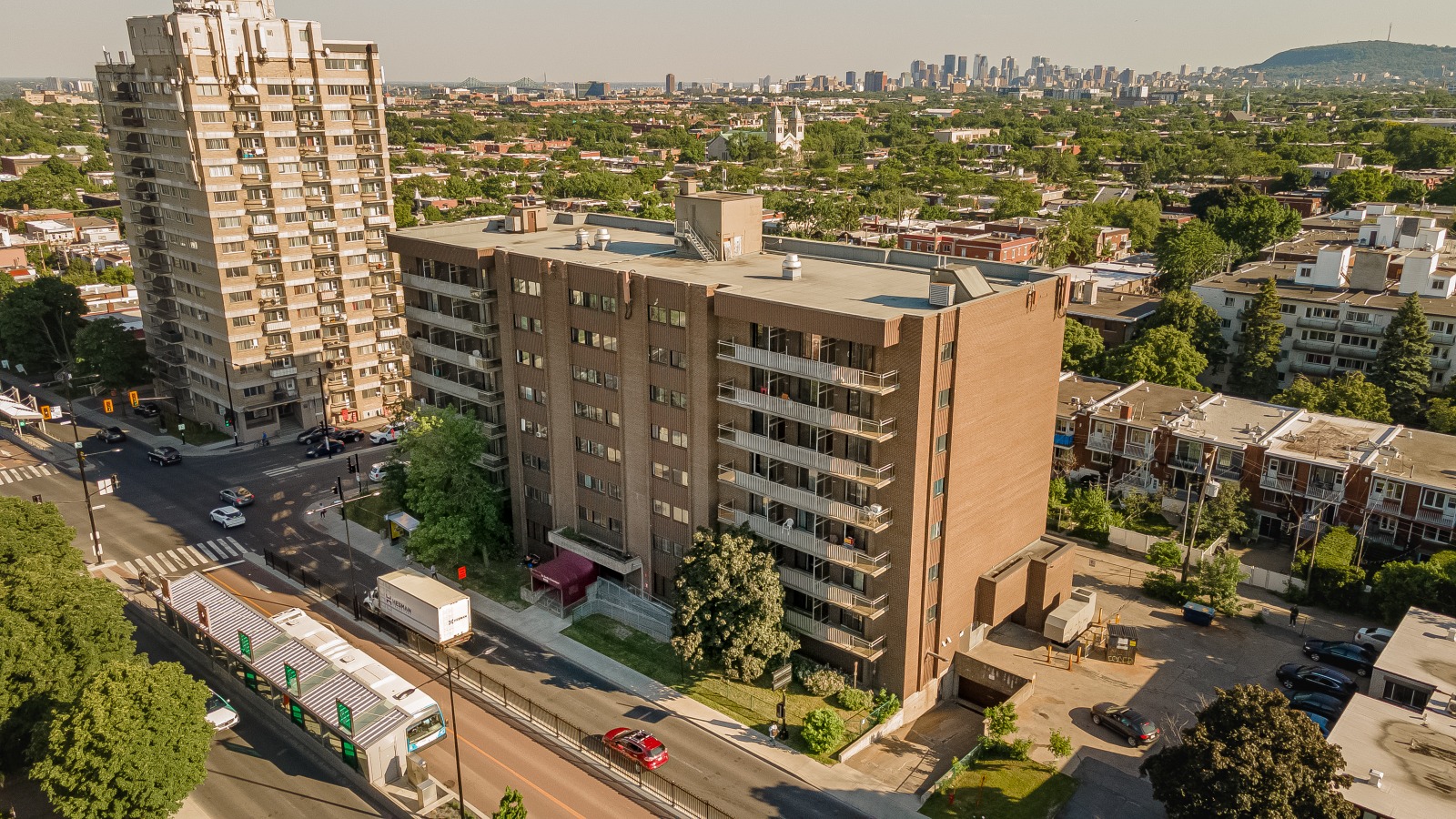 Aerial view of urban neighborhood with apartment buildings, streets, greenery, and a city skyline in the background.