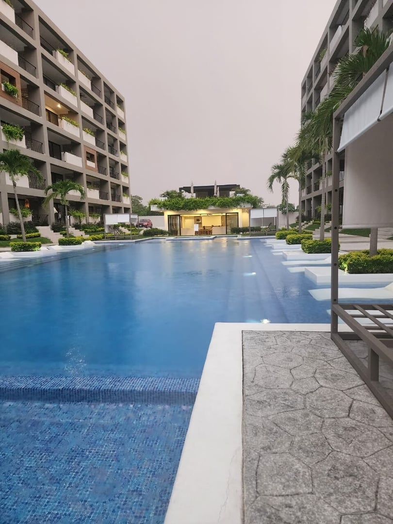 Modern apartment complex with a large swimming pool surrounded by landscaped greenery and tall buildings.