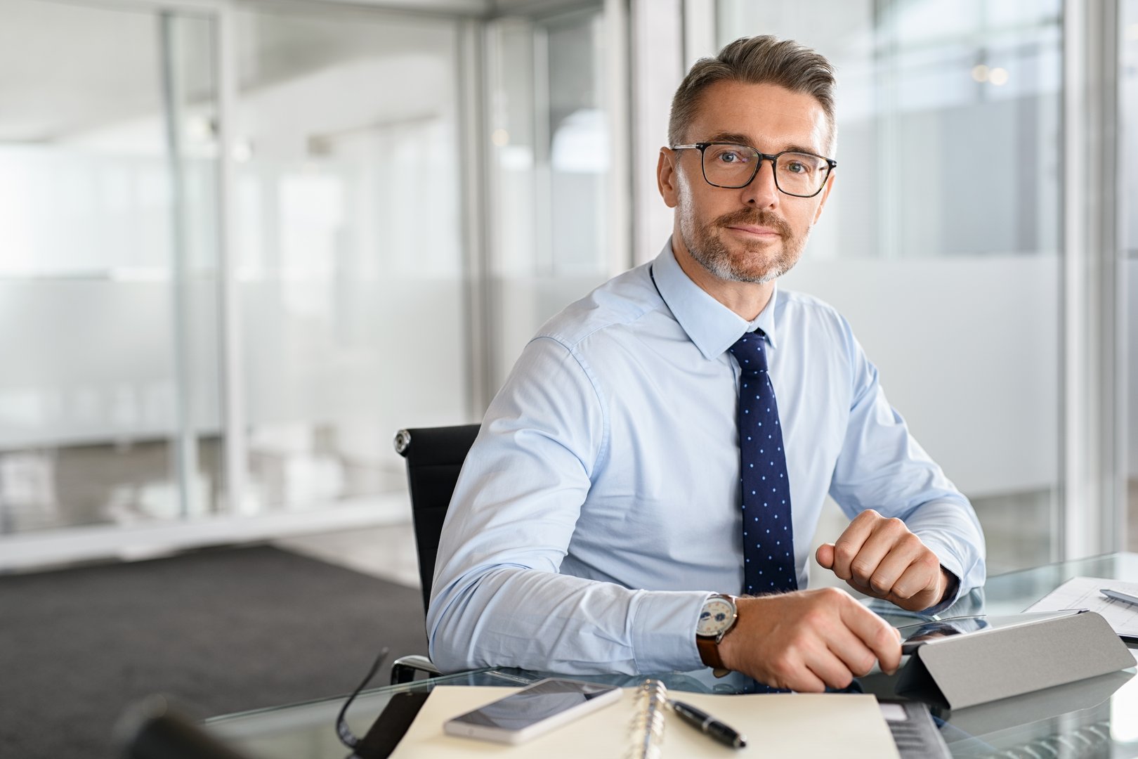 Portrait of mature businessman using digital tablet at work. Successful business entrepreneur working in a modern office. Satisfied mid adult manager sitting at desk and checking email while looking at camera with copy space.
