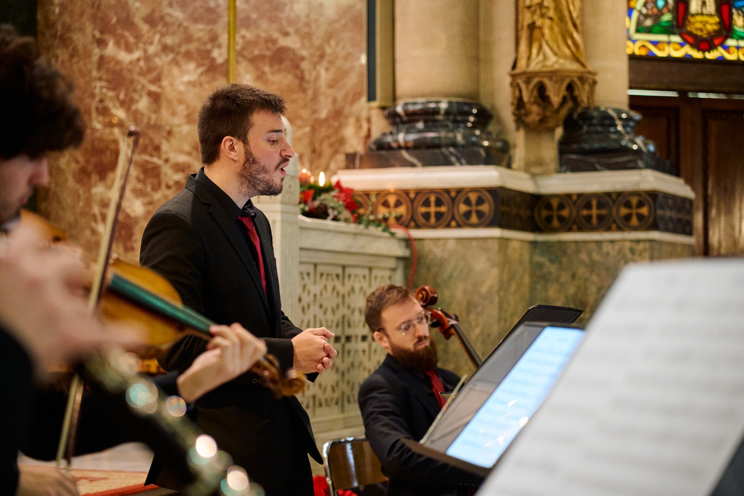 Musicians perform in a church, showcasing instruments such as violin and cello, while a vocalist leads the song. The setting provides a serene atmosphere, enhancing the music experience.