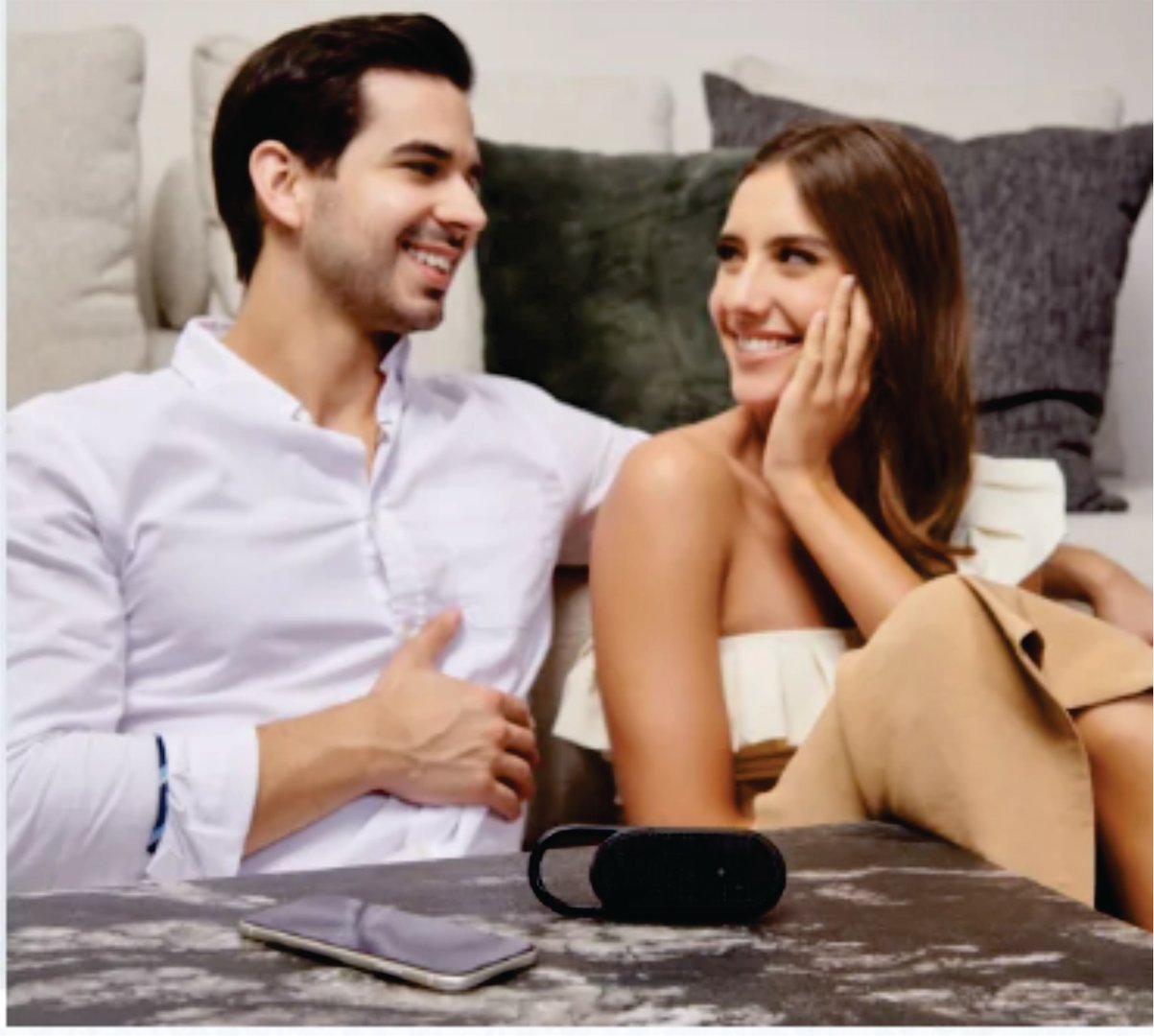A couple sitting on a couch, smiling at each other. A smartphone and a small speaker are on the table in front of them.
