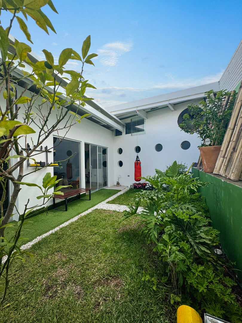 Small backyard with green lawn, punching bag, potted plants, and modern white building with circular windows.