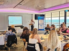 A speaker presents at a conference room with attendees seated at tables, a projection screen, and banners in the background.