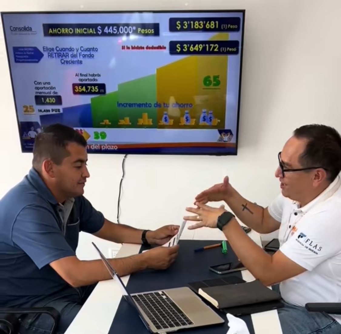 Two men discussing financial information at a table with a laptop, in front of a screen displaying savings details.