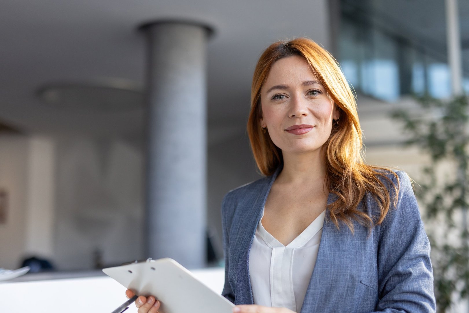 A confident young businesswoman stands in an office, holding a clipboard and smiling, ready to take on new challenges in her professional journey.
