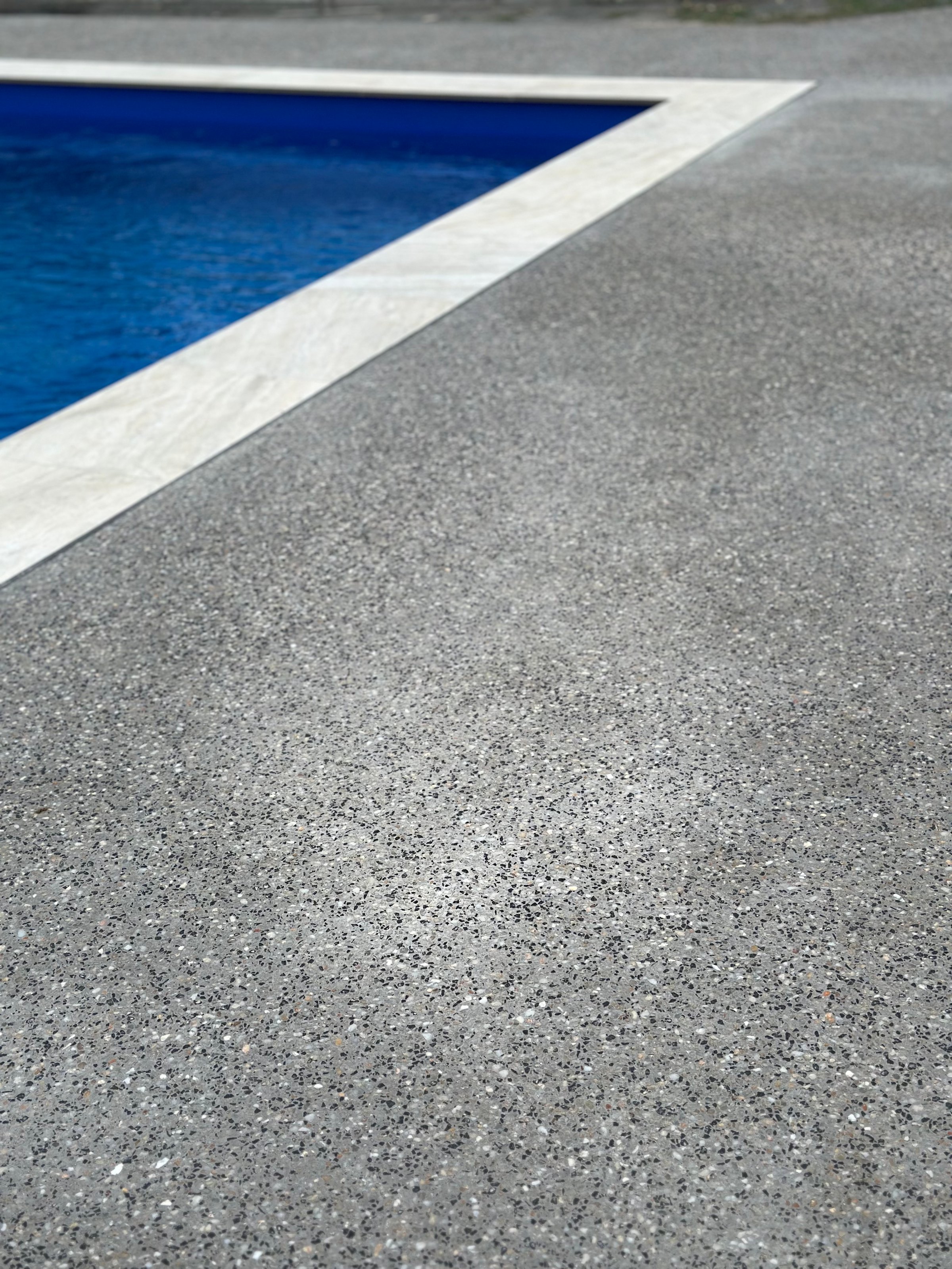 Close-up of a textured concrete pool deck edge leading to a blue swimming pool with water ripples.