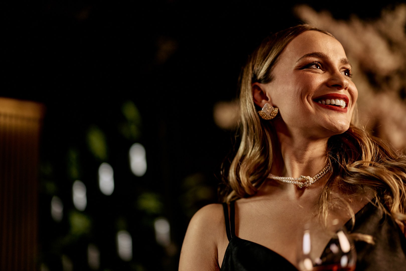 A smiling woman in an elegant setting wearing jewelry, holding a glass of wine.