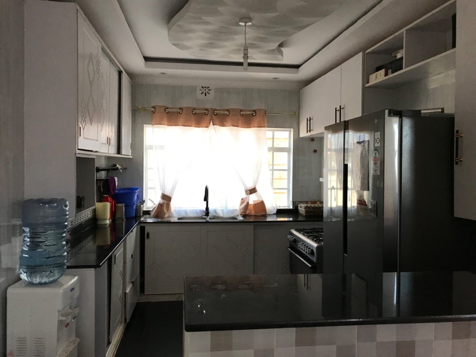 Modern kitchen with grey cabinets, stainless steel fridge, water dispenser, and a window with sheer curtains.